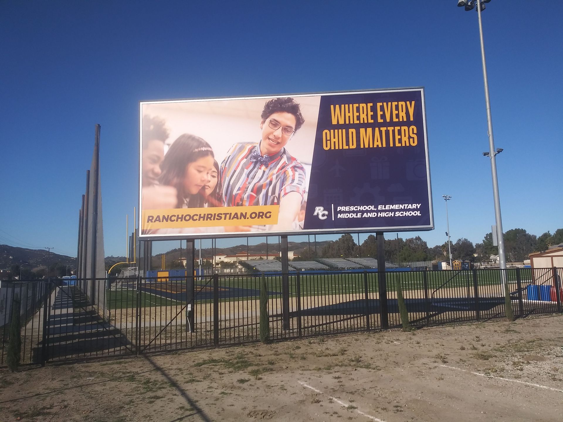 Billboard in a fenced field with a mother and child, reading “Where Every Child Matters” against a blue sky