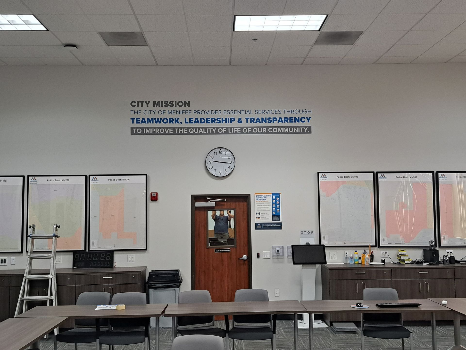 Community room with tables and chairs, bulletin boards, wall clock, and a sign about teamwork and leadership.