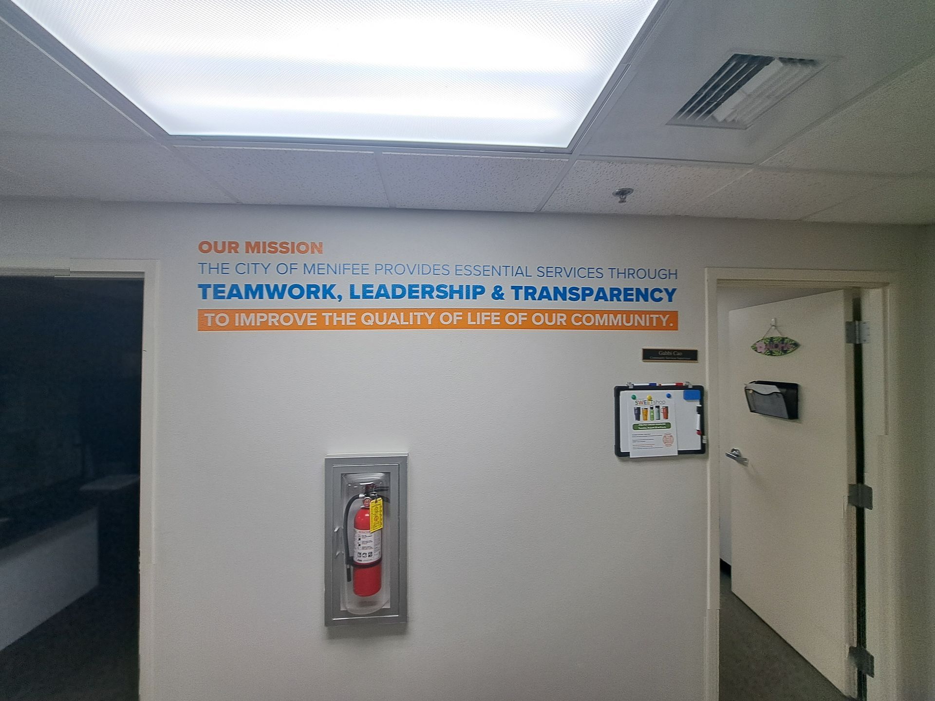 Office hallway with wall sign reading “TEAMWORK, LEADERSHIP & TRANSPARENCY” and a fire extinguisher cabinet