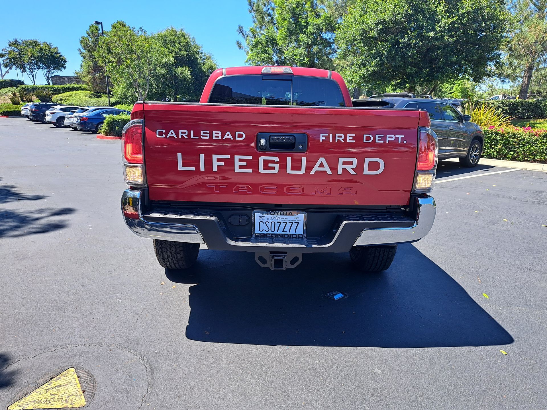 Red lifeguard pickup truck parked on a sunny road with trees and parked cars nearby