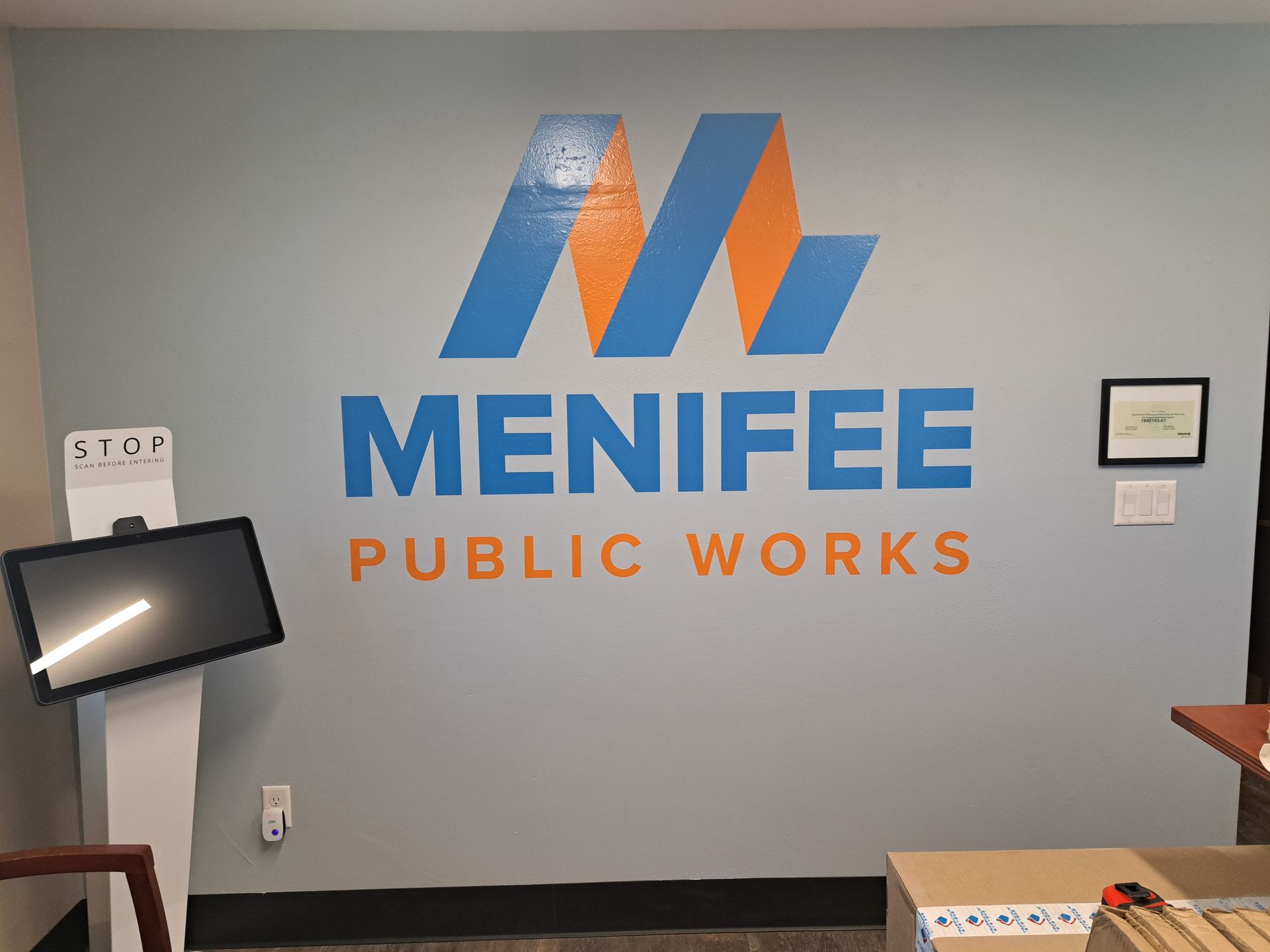 Menifee Public Works logo on a gray office wall with blue and orange lettering
