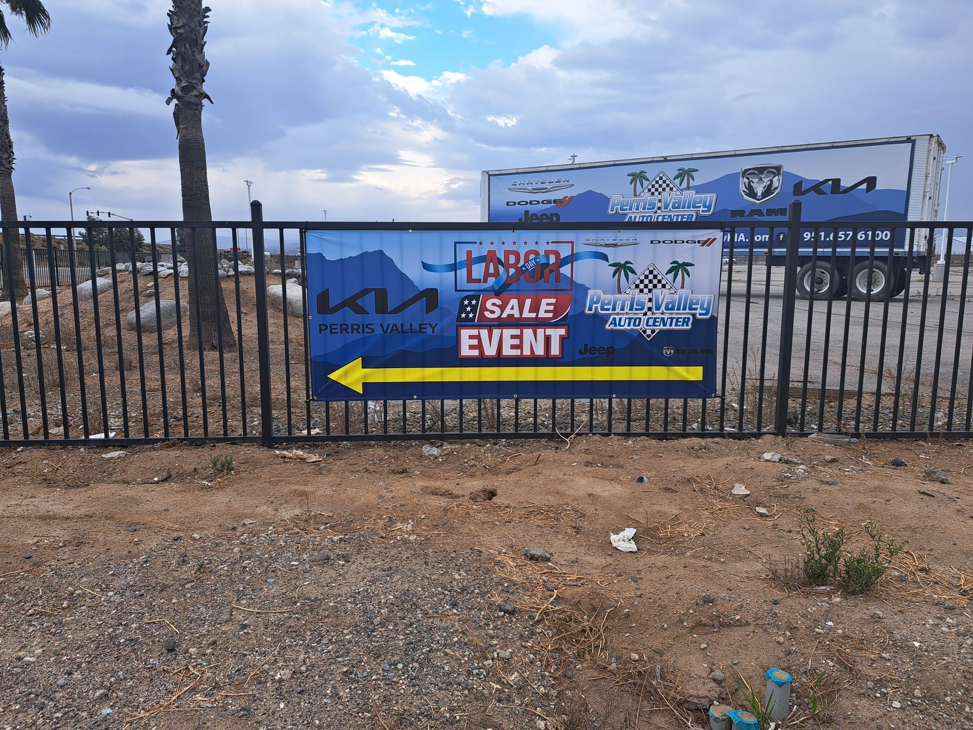 Banner on a fence advertising a blue truck sale event in a dusty lot, with a truck trailer in the background
