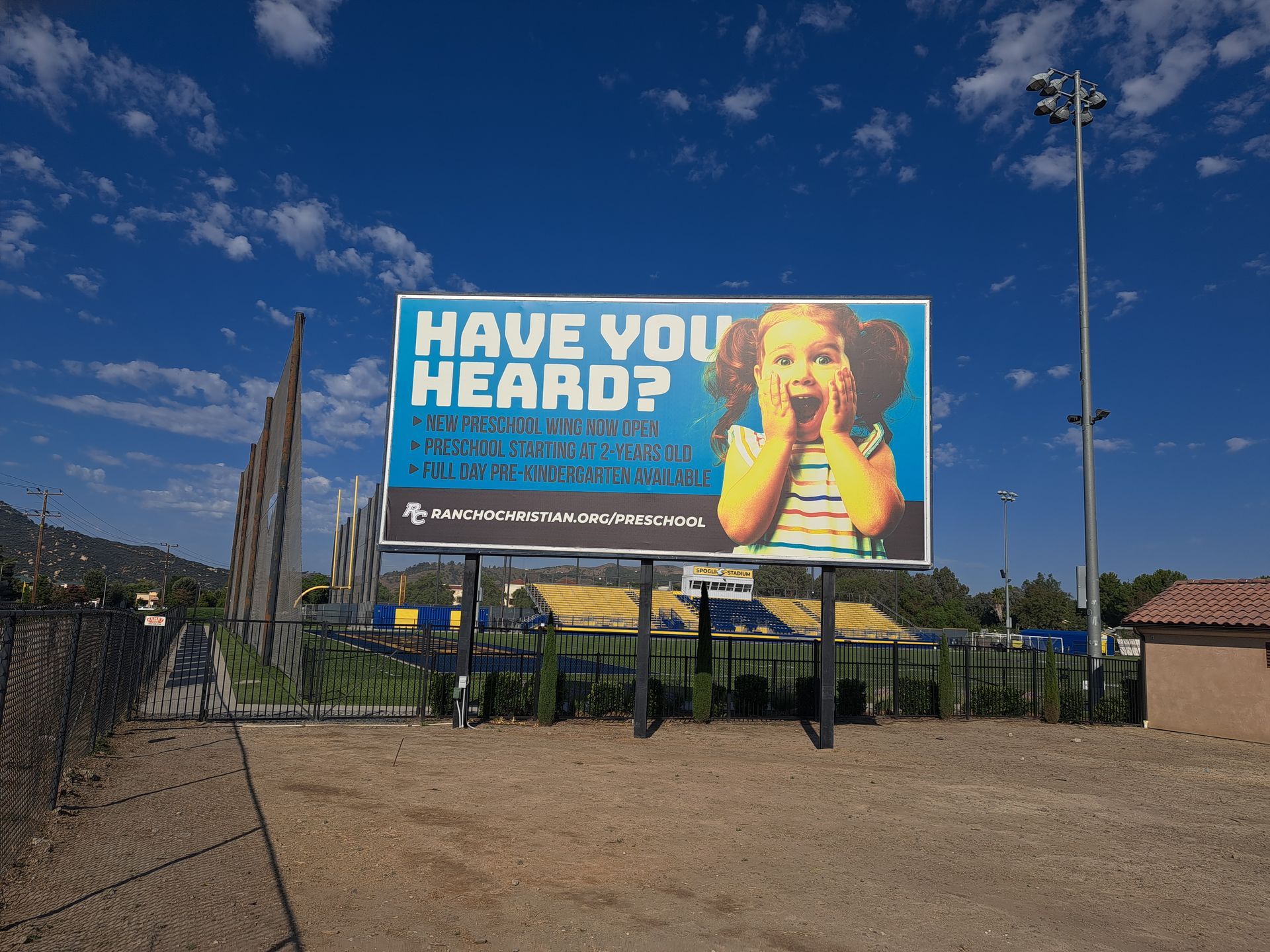 Billboard in a gravel lot under a blue sky, reading “HAVE YOU HEARD?” with a person holding a megaphone.