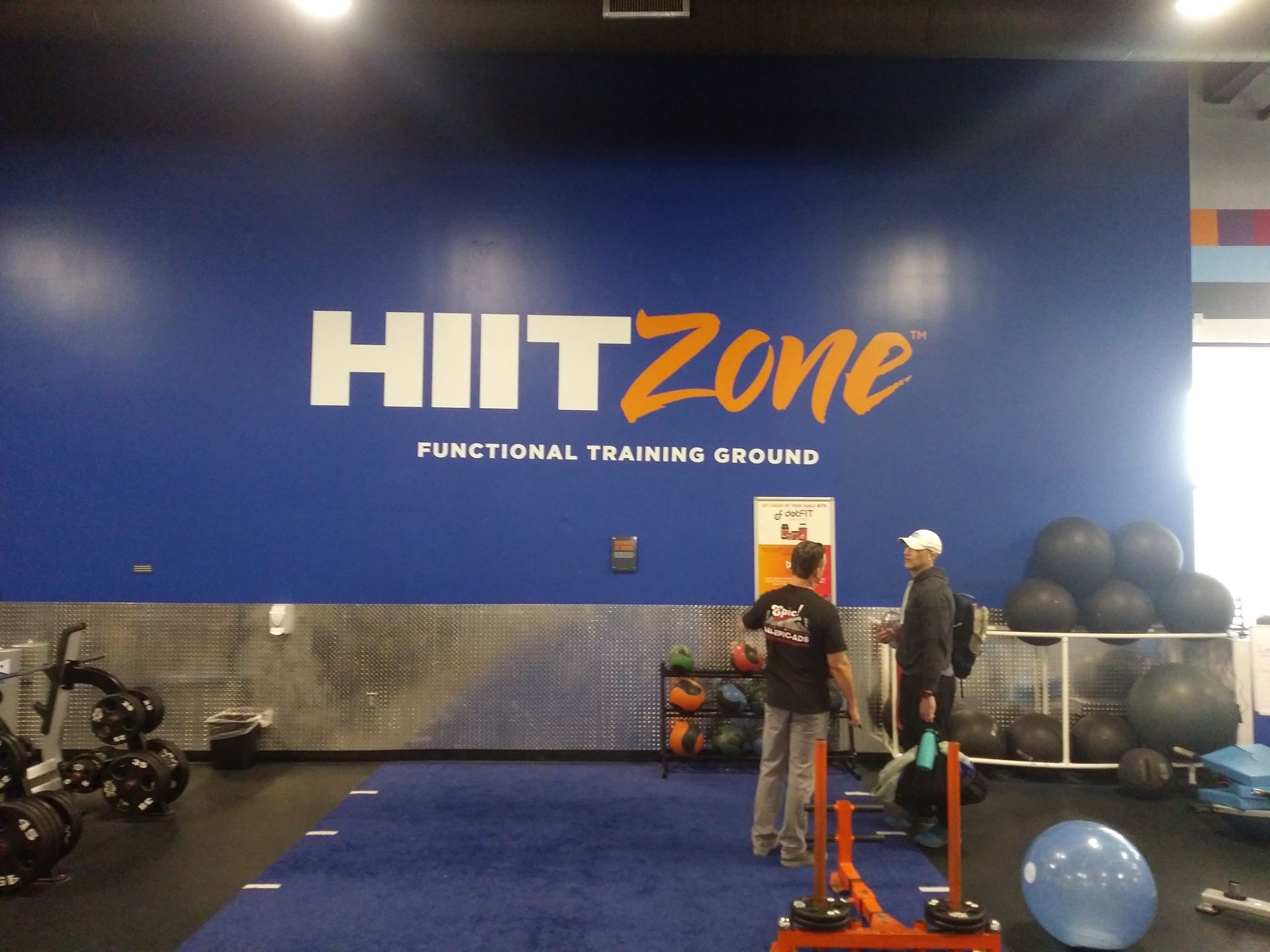 Two people training in a HIITZone gym with blue walls, equipment, and exercise balls.