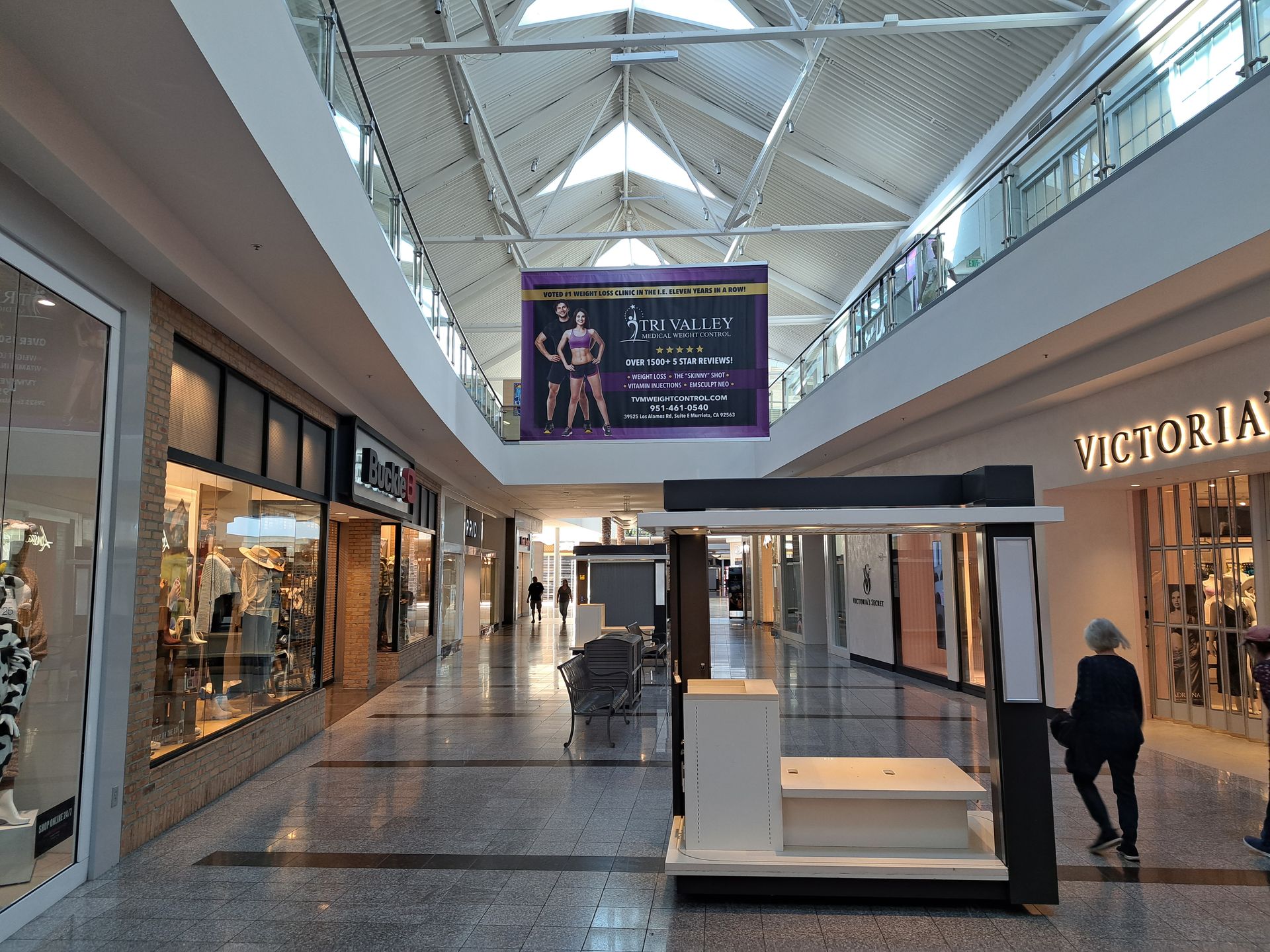 Bright shopping mall corridor with glass storefronts, a central kiosk, and a large hanging ad display.