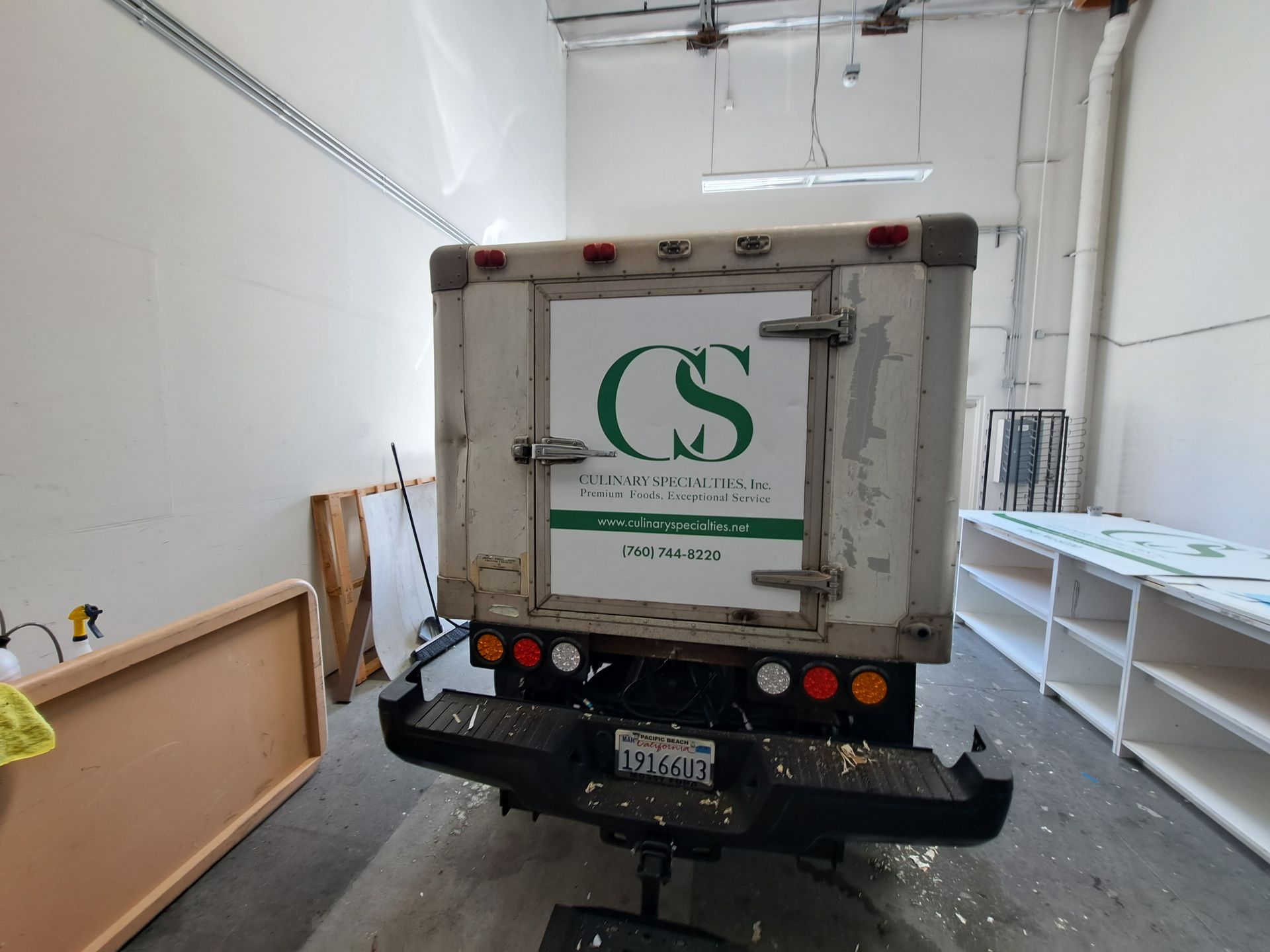White box truck parked inside a narrow loading bay beside stacked wooden panels and white shelving units.