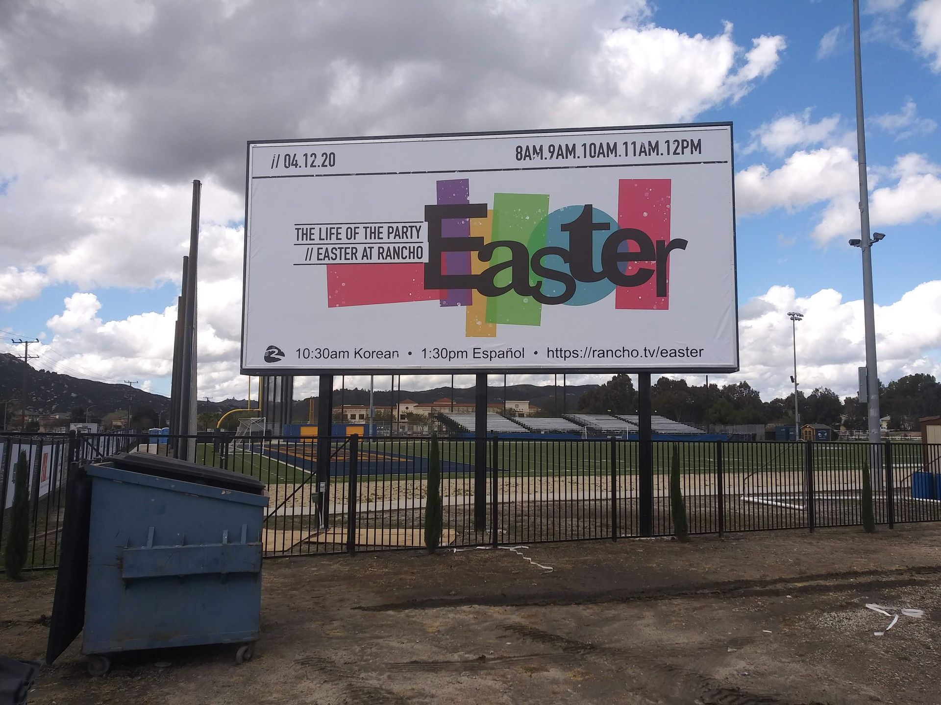 Large “Easter” billboard at a sports field, with a blue dumpster in front and cloudy sky overhead