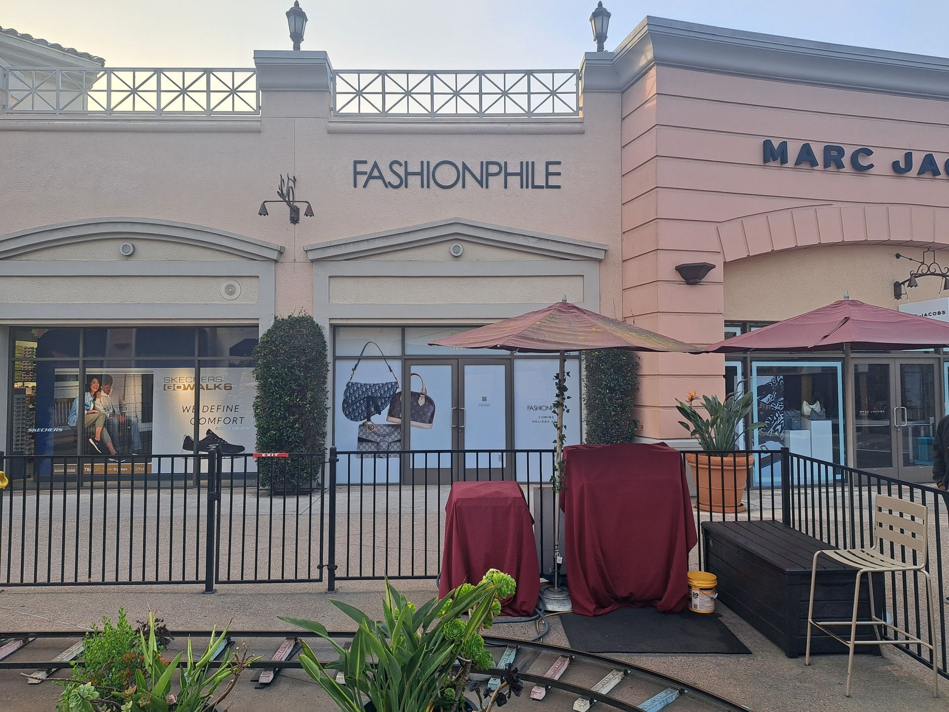 Storefronts with a Fashionphile sign, outdoor seating, and covered tables on a sidewalk mall.