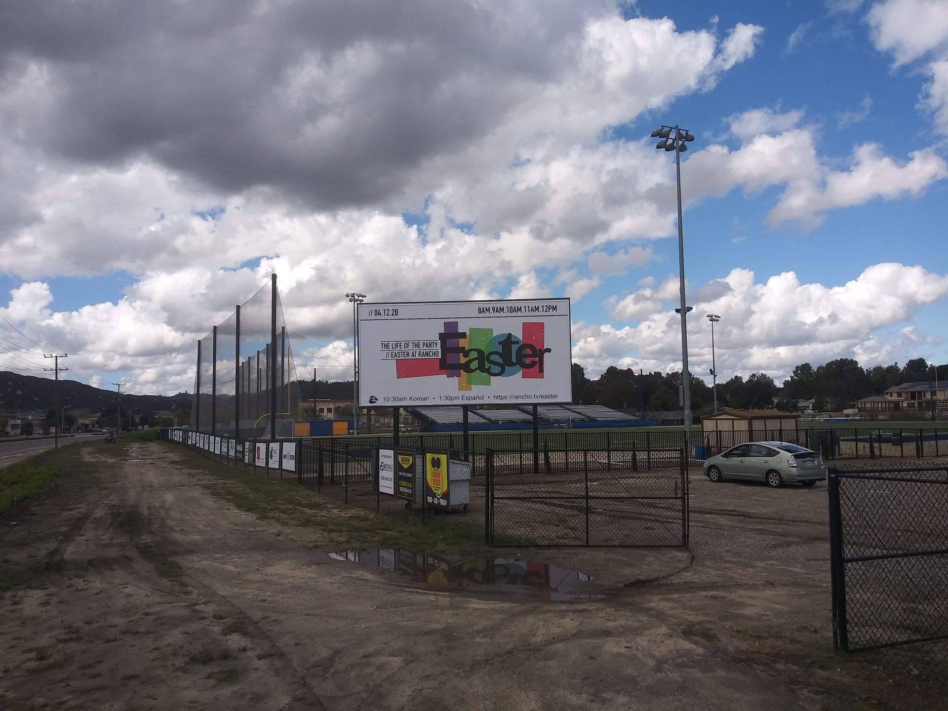 Outdoor dirt lot with a fence, parked cars, and a large colorful billboard under a cloudy sky.