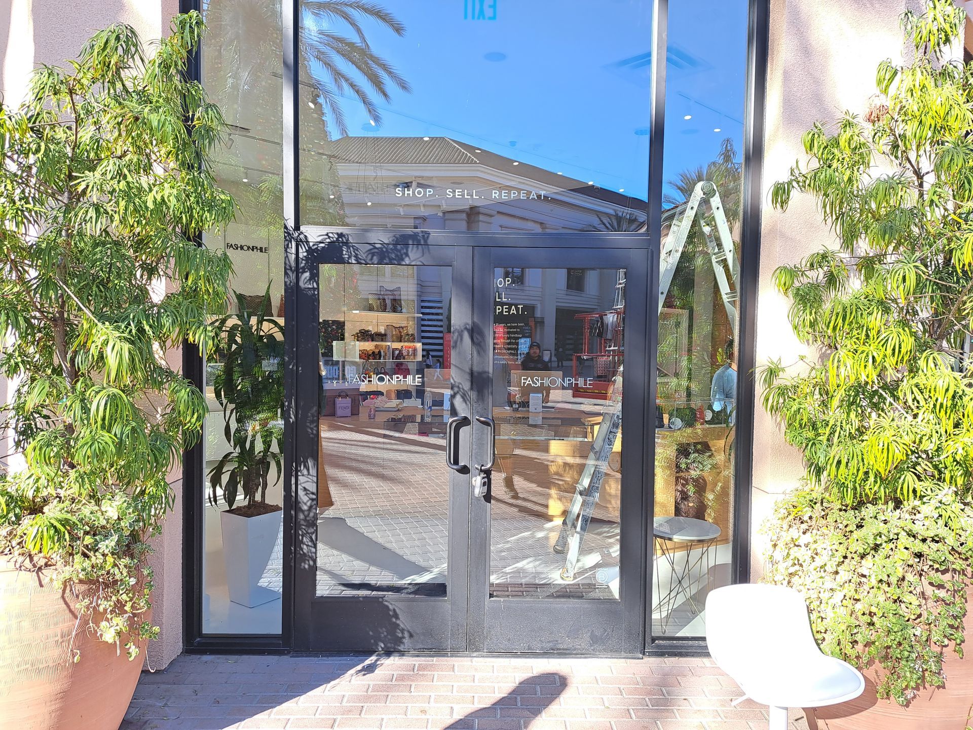 Glass entrance to a building with potted trees, a patio, and a white chair outside