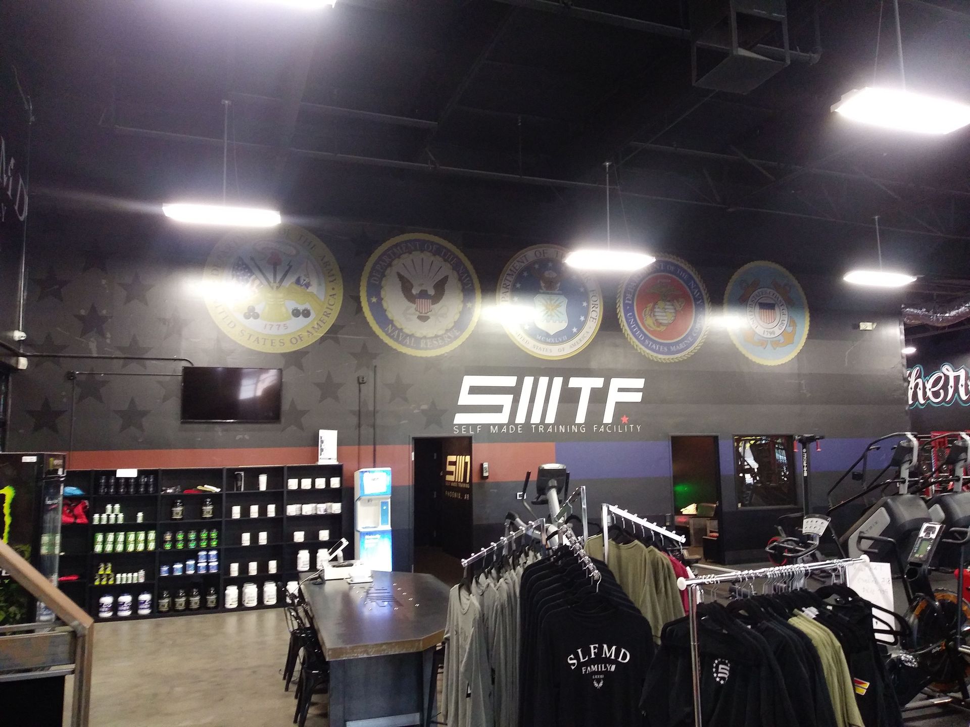 Indoor gym pro shop with racks of clothing, shelves of supplements, and large wall logos under bright lights