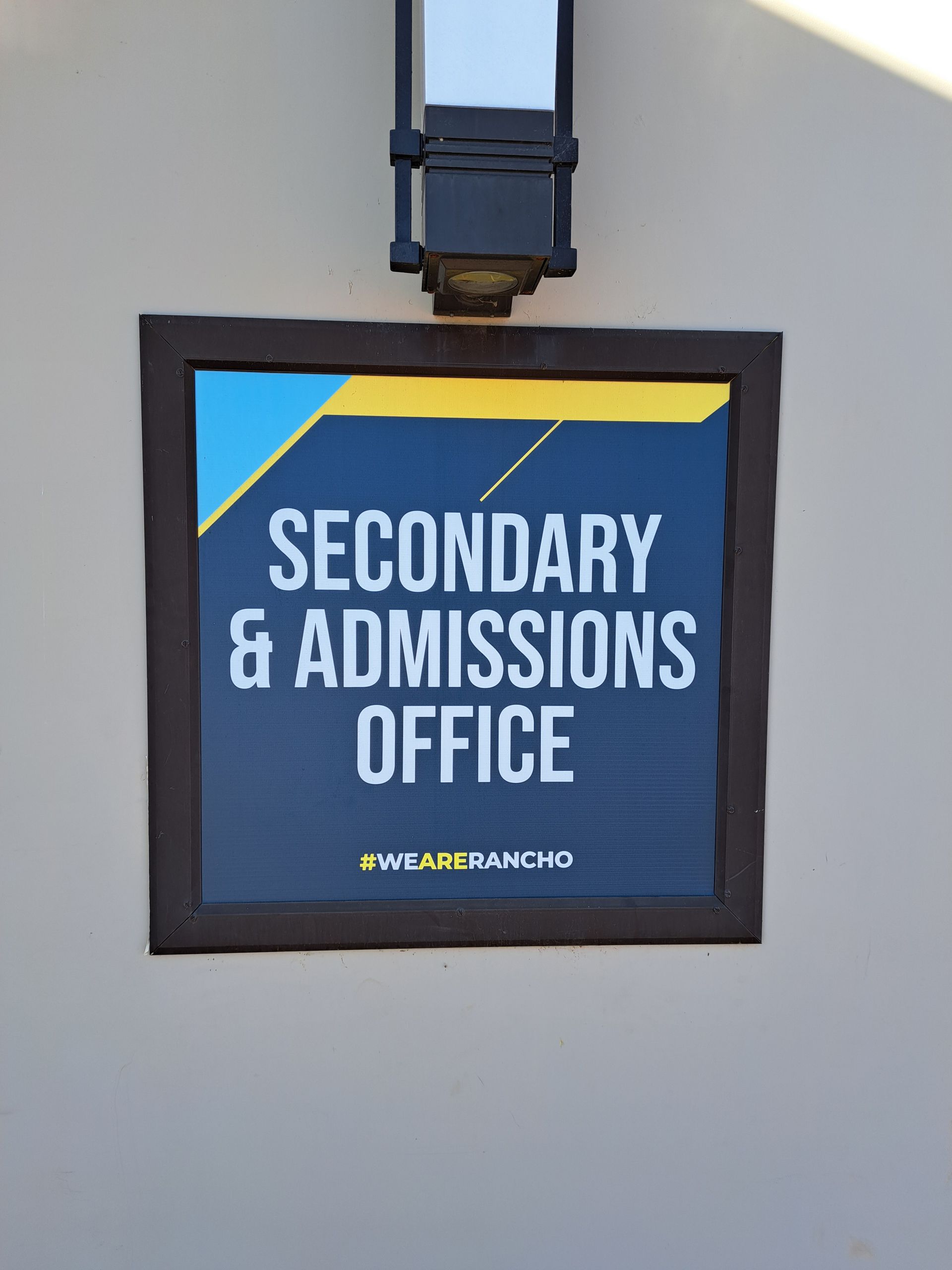 Sign for Secondary & Admissions Office on a wall, in blue and yellow with a black frame