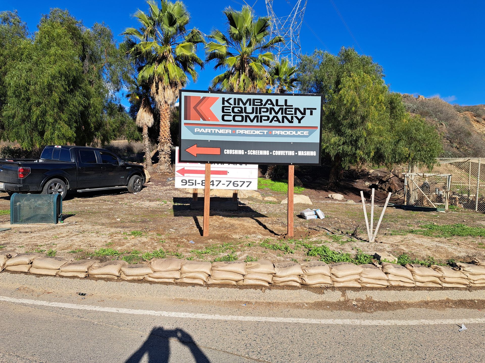 Kimball Bay Recovery sign beside a road, with palm trees, dry landscaping, and blue sky.