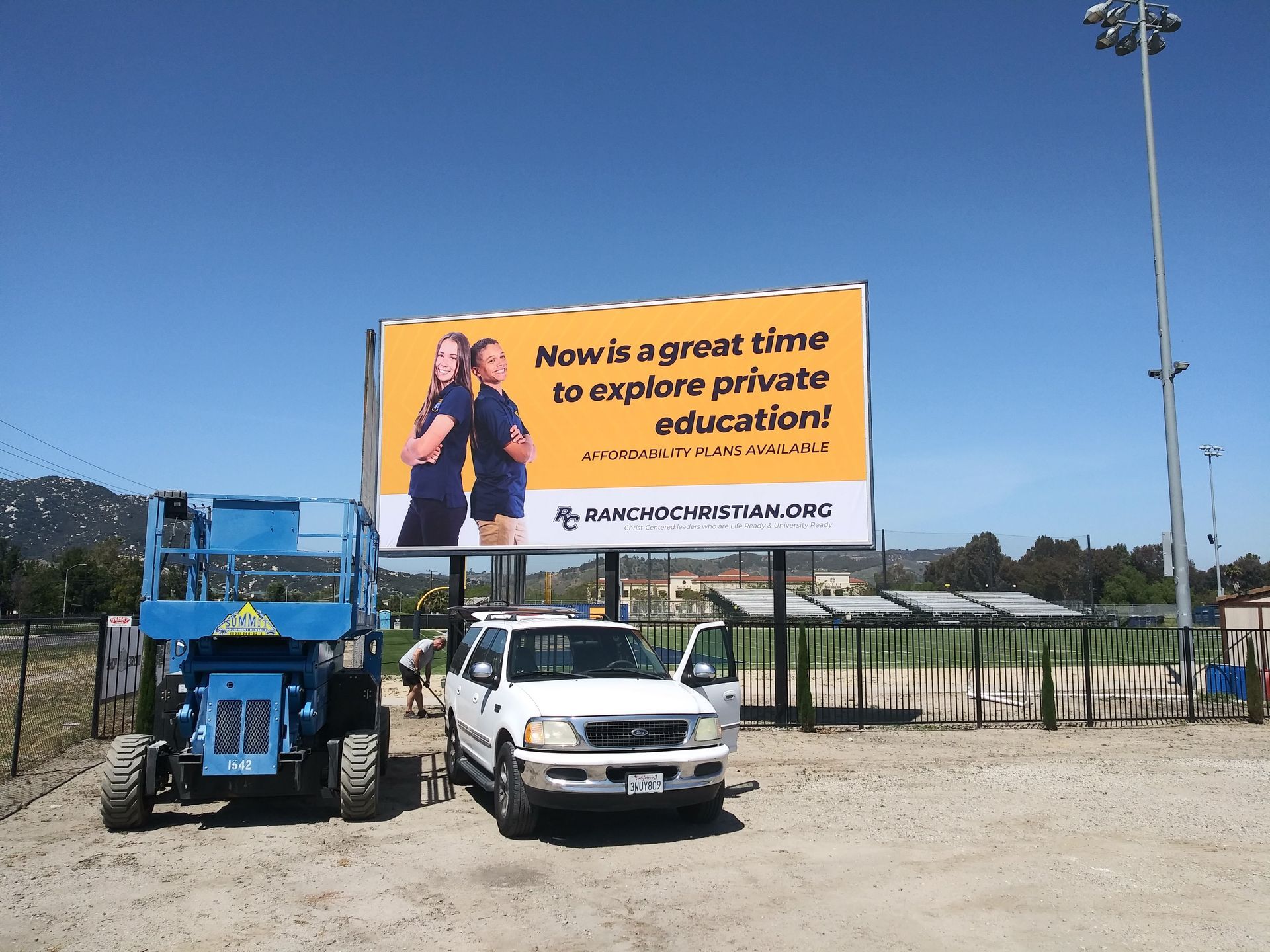 Billboard for “Now is a great time to explore private education” beside a blue lift and parked white car