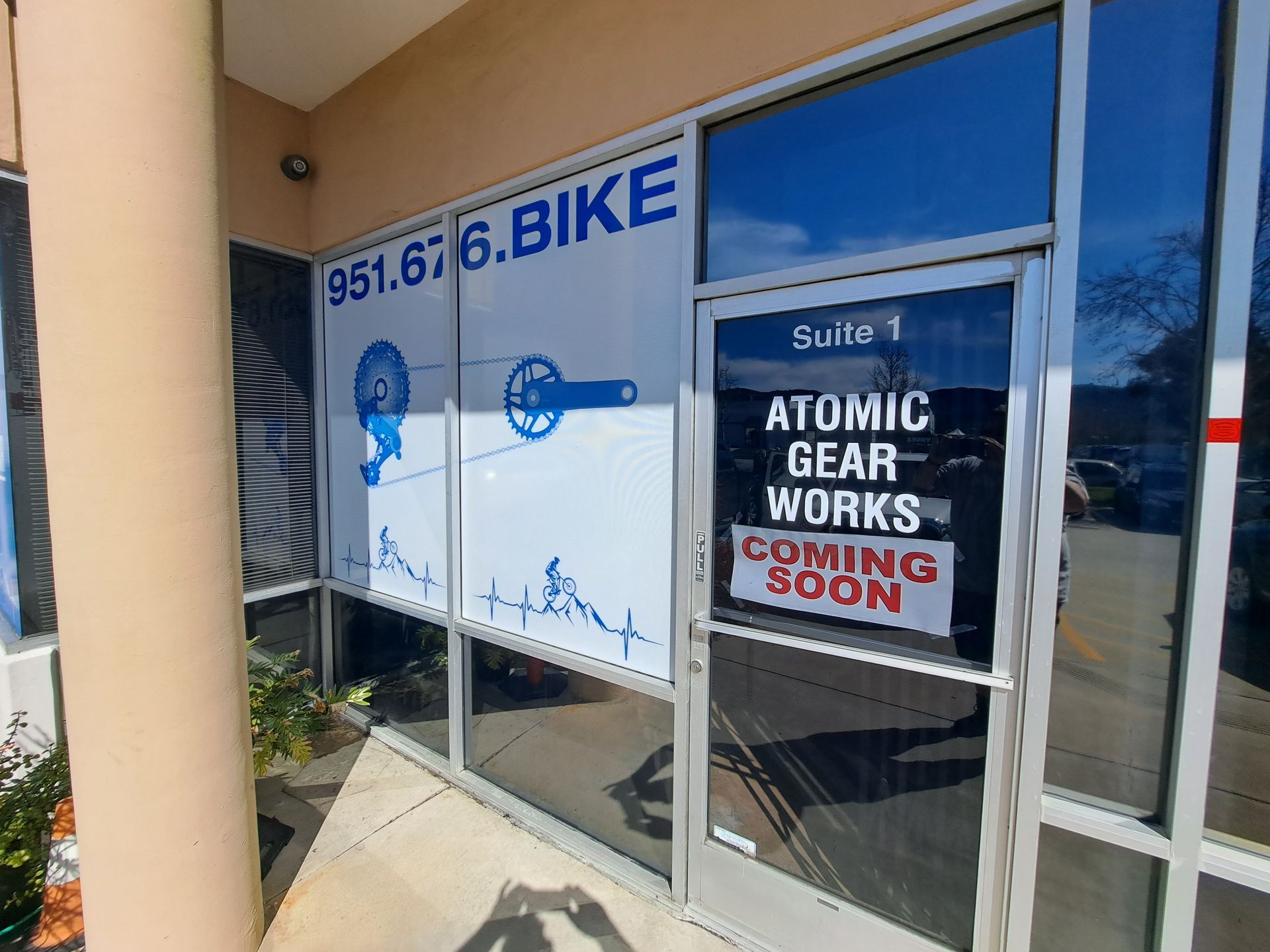 Storefront windows with blue “Bike” sign and a “Atomic Gear Works coming soon” notice