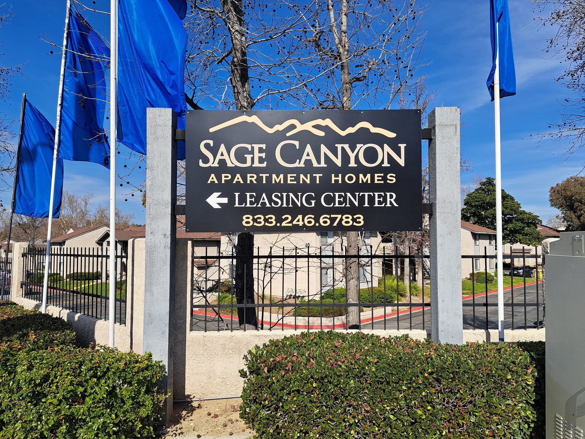 Sage Canyon Apartment Homes leasing center sign with blue flags and landscaped entrance