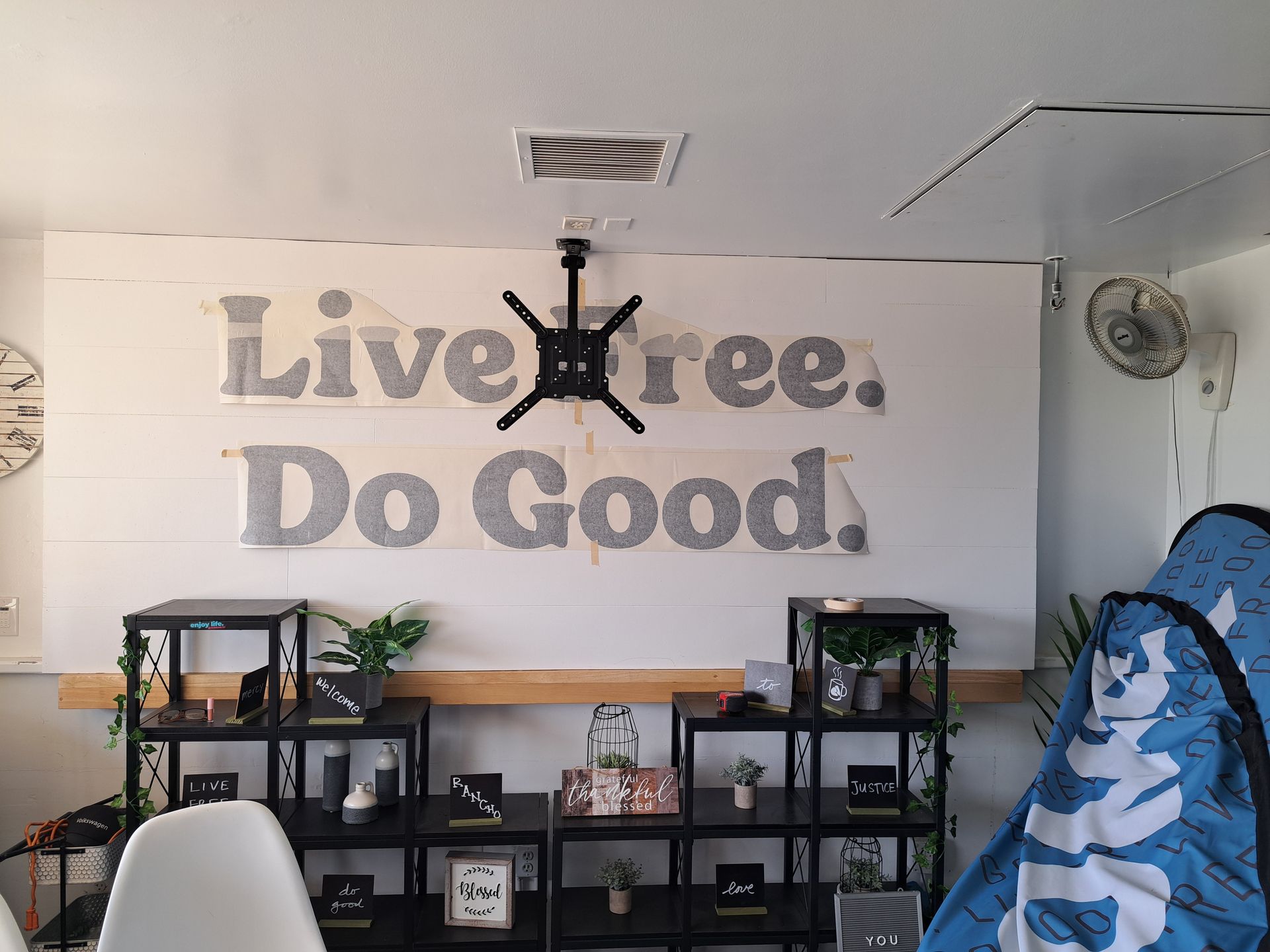 Office wall with “Live Free. Do Good.” sign above shelves, fan, and plants