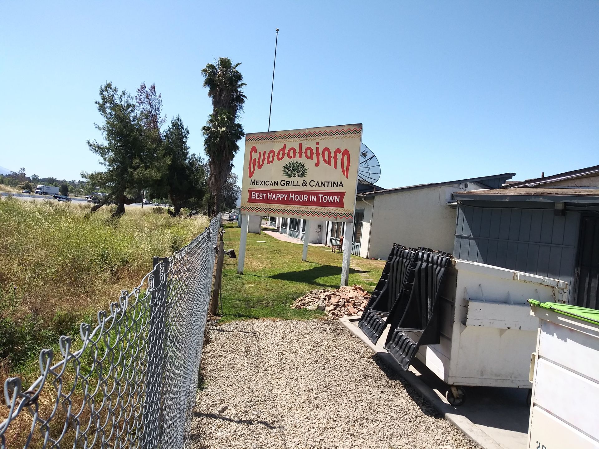 Roadside sign in a gravel path beside a fence, with trees and a building under a clear blue sky.