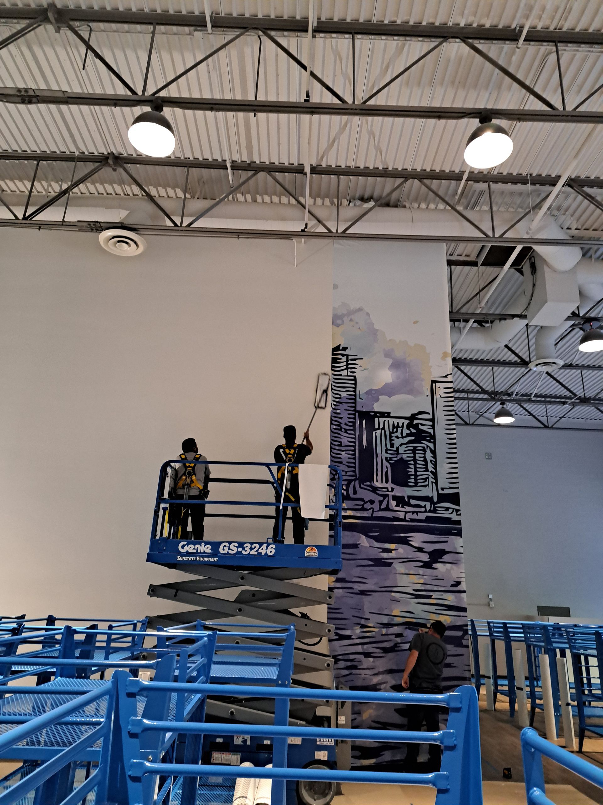 People on a blue lift inside a large indoor industrial space with high ceilings and mural wall