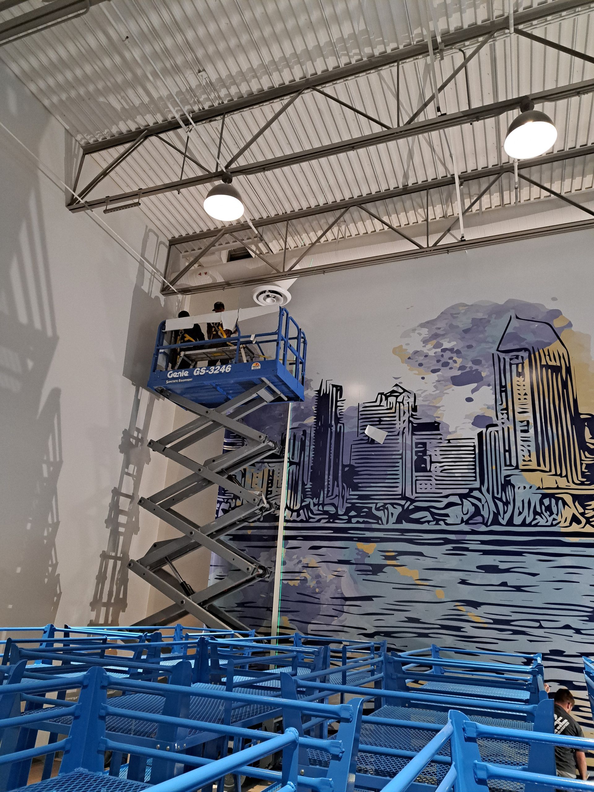 Blue scissor lift with worker painting a large mural on a warehouse wall under high ceiling lights