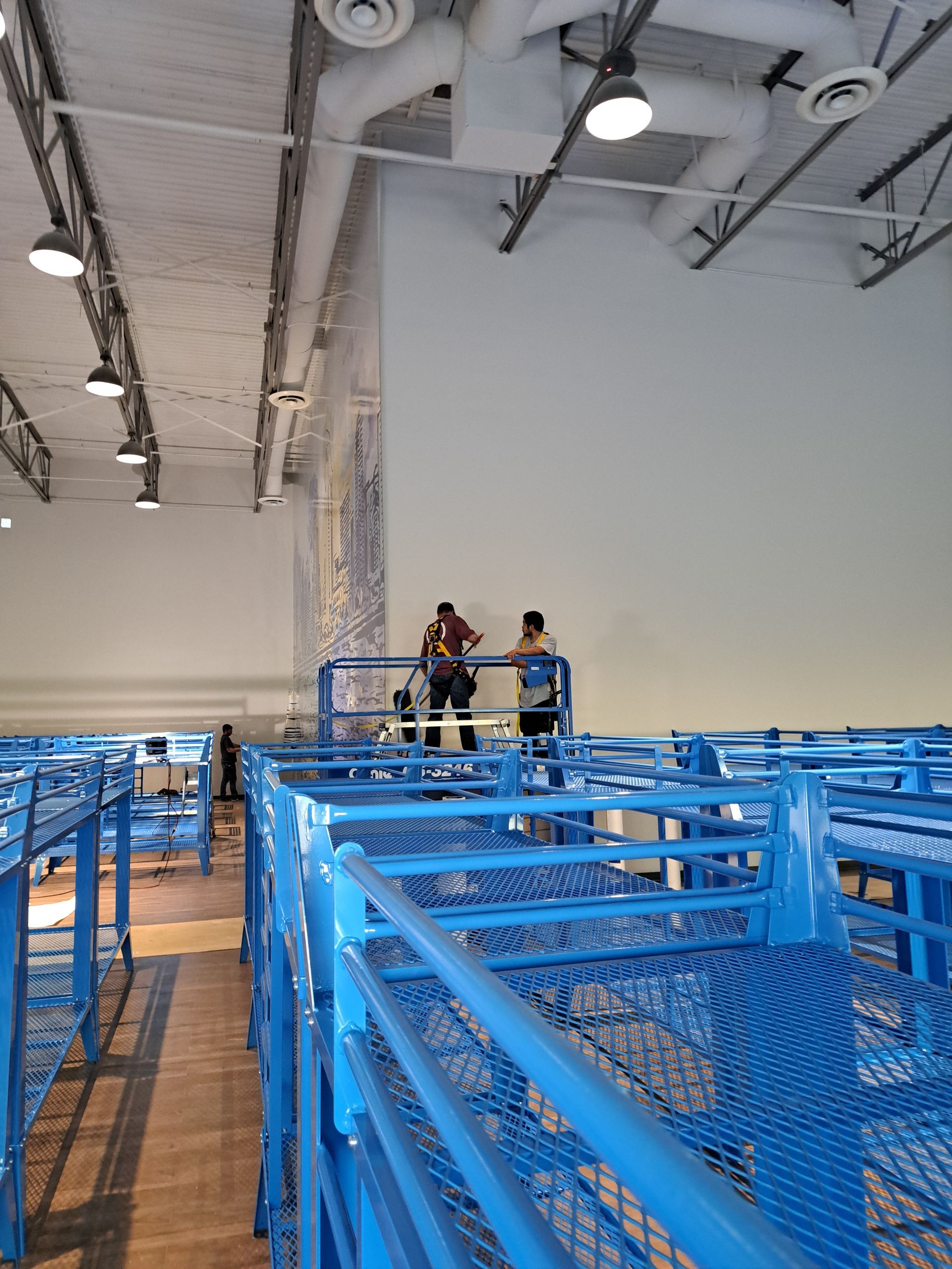 Two people standing on a yellow platform above rows of blue metal racks in a large industrial warehouse