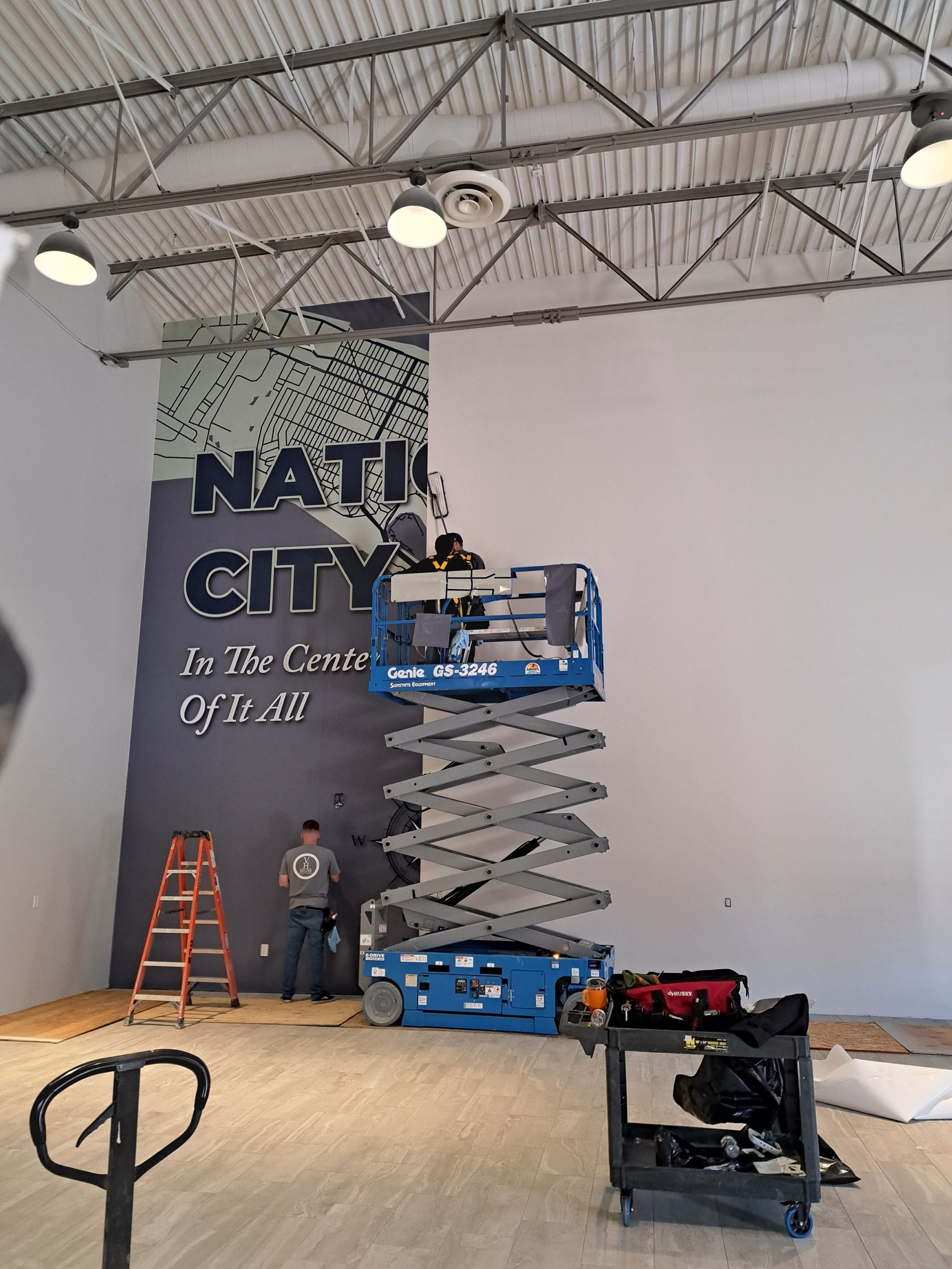 Workers on ladders and a scissor lift painting a wall mural inside a large warehouse space.