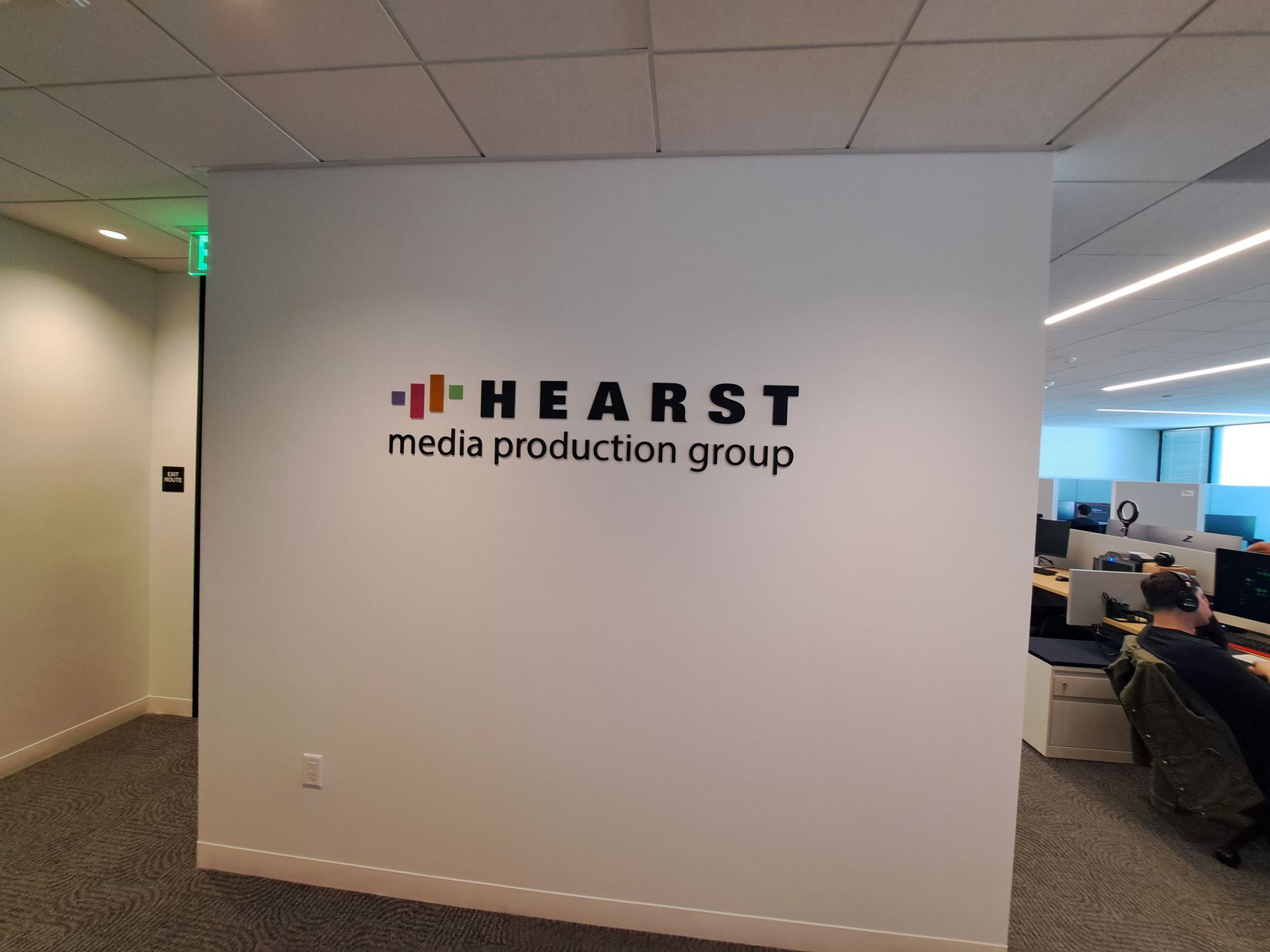 HEARST media production group sign on an office wall, with desks and a seated person in the background