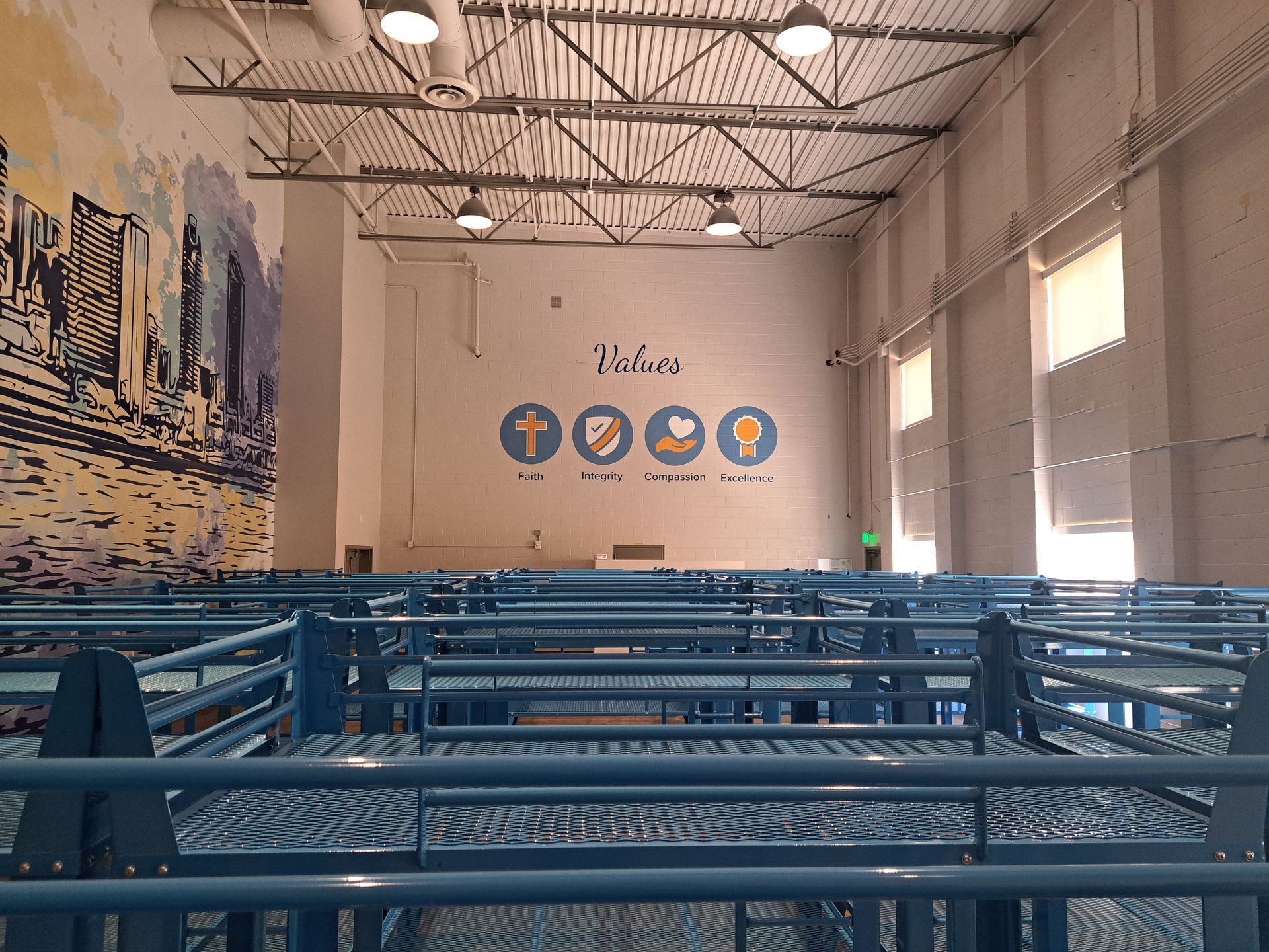 Empty blue metal benches in a bright indoor hall with a stage and mural on the far wall.