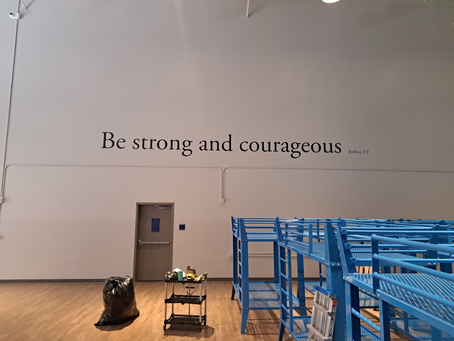 Gymnasium wall with “Be strong and courageous …” text, blue bleachers, and a lone bag on the floor.