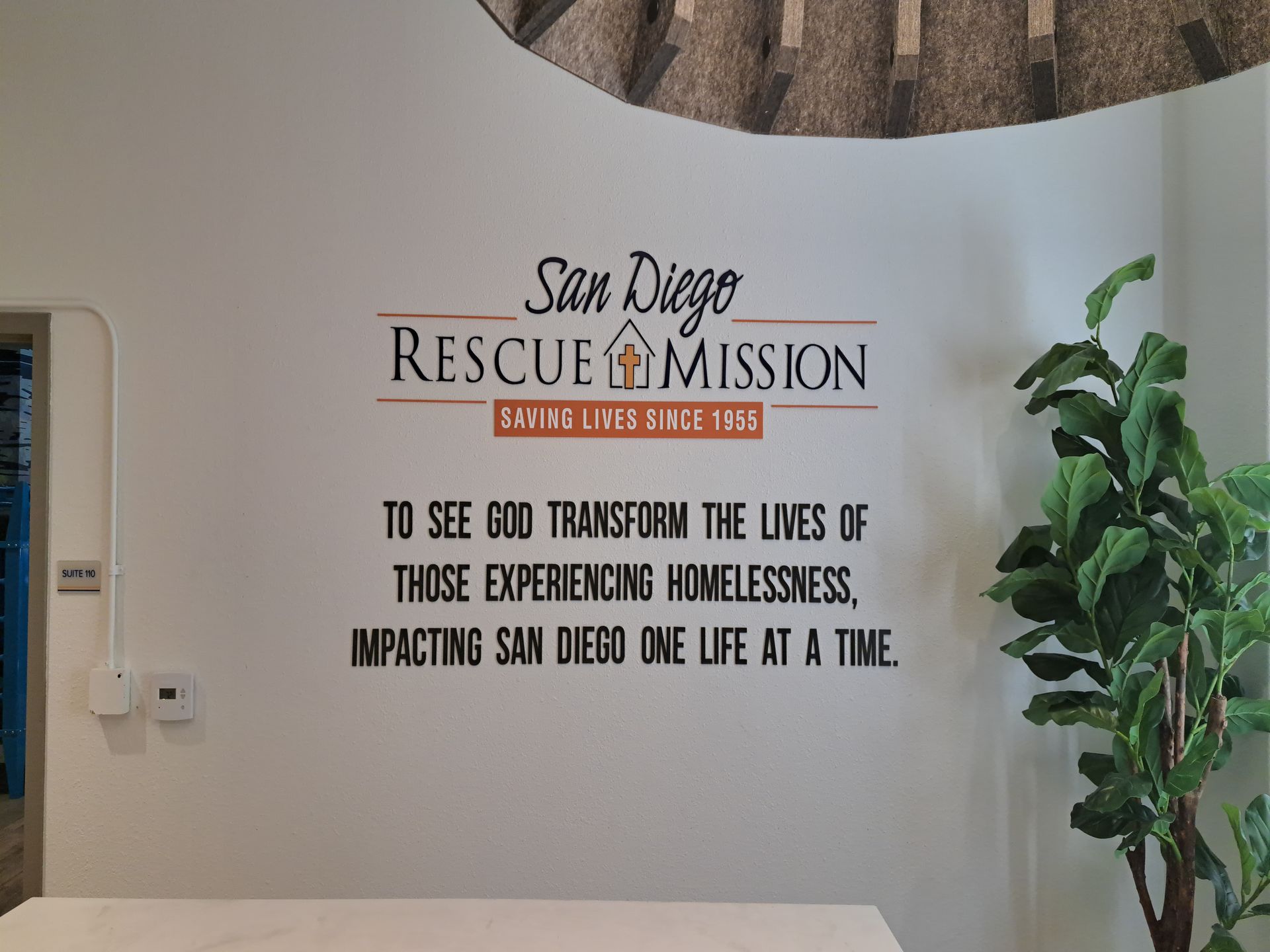 San Diego Rescue Mission wall sign with mission statement in black lettering beside a potted plant