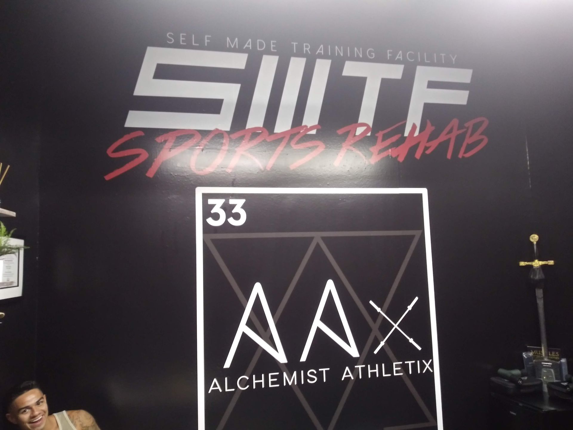 SWTF Sports Rehab sign and Alchemist Athletics logo on a black wall, with gym equipment nearby.