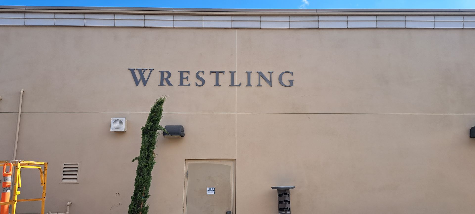Wrestling sign on a beige building wall with a door and climbing vine nearby