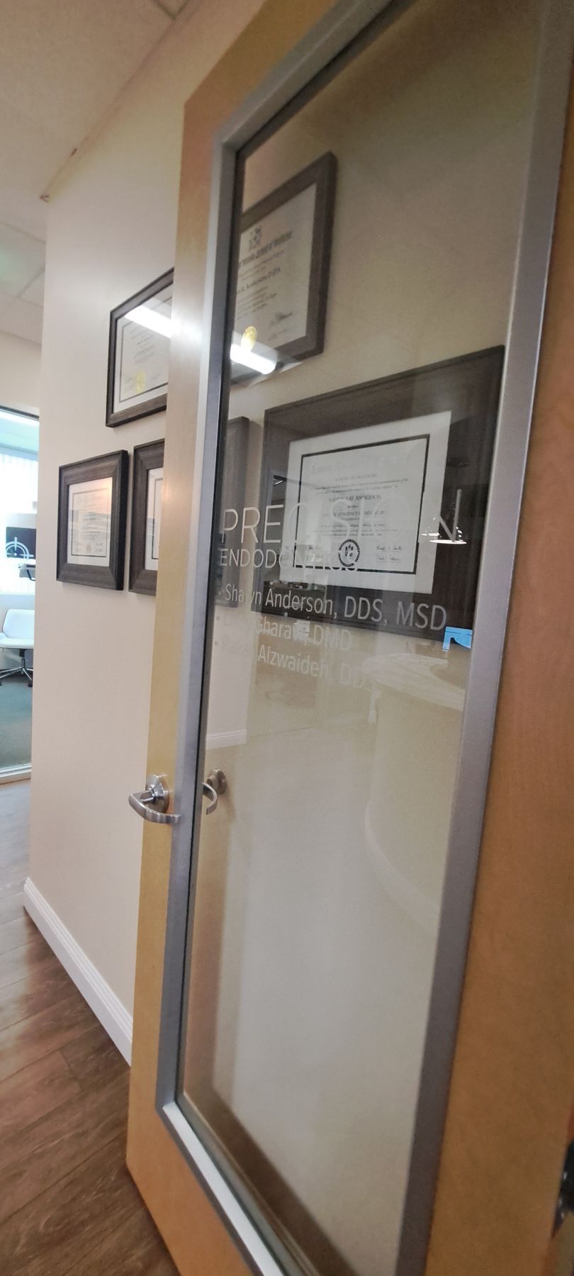 Hallway with framed certificates on a wall beside a glass office door and wooden trim