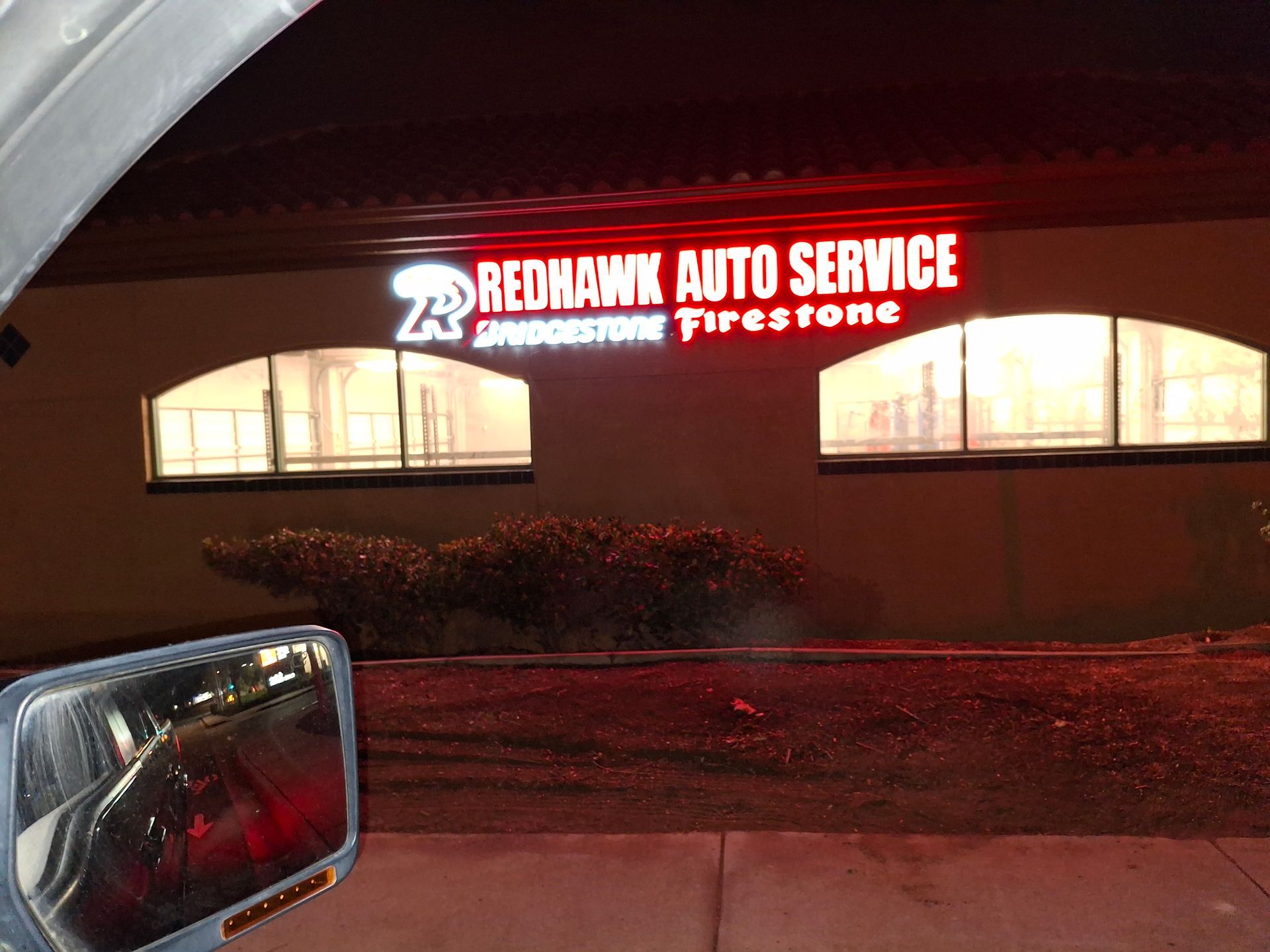 Redman Auto Service sign glowing red at night above a storefront with lit windows and a car mirror in foreground
