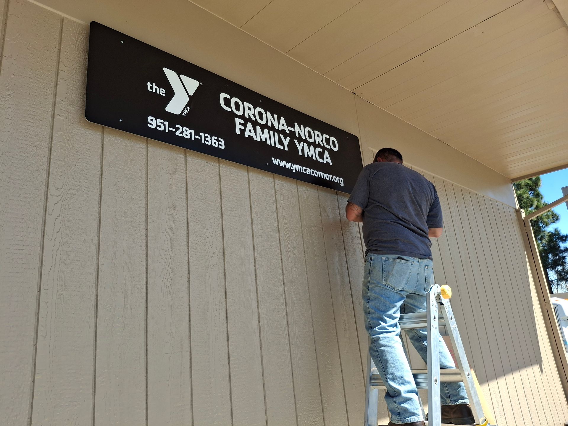 Worker on a ladder installing a black YMCA sign on a beige building wall.