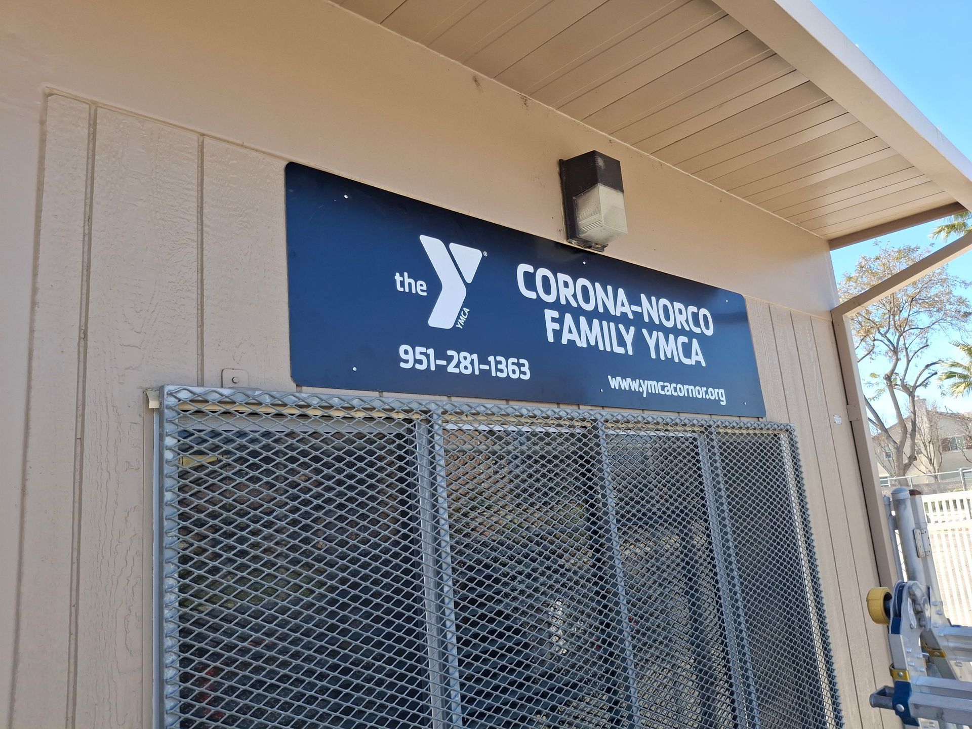 Corona-Norco Family YMCA sign above a chain-link gate on a beige building exterior