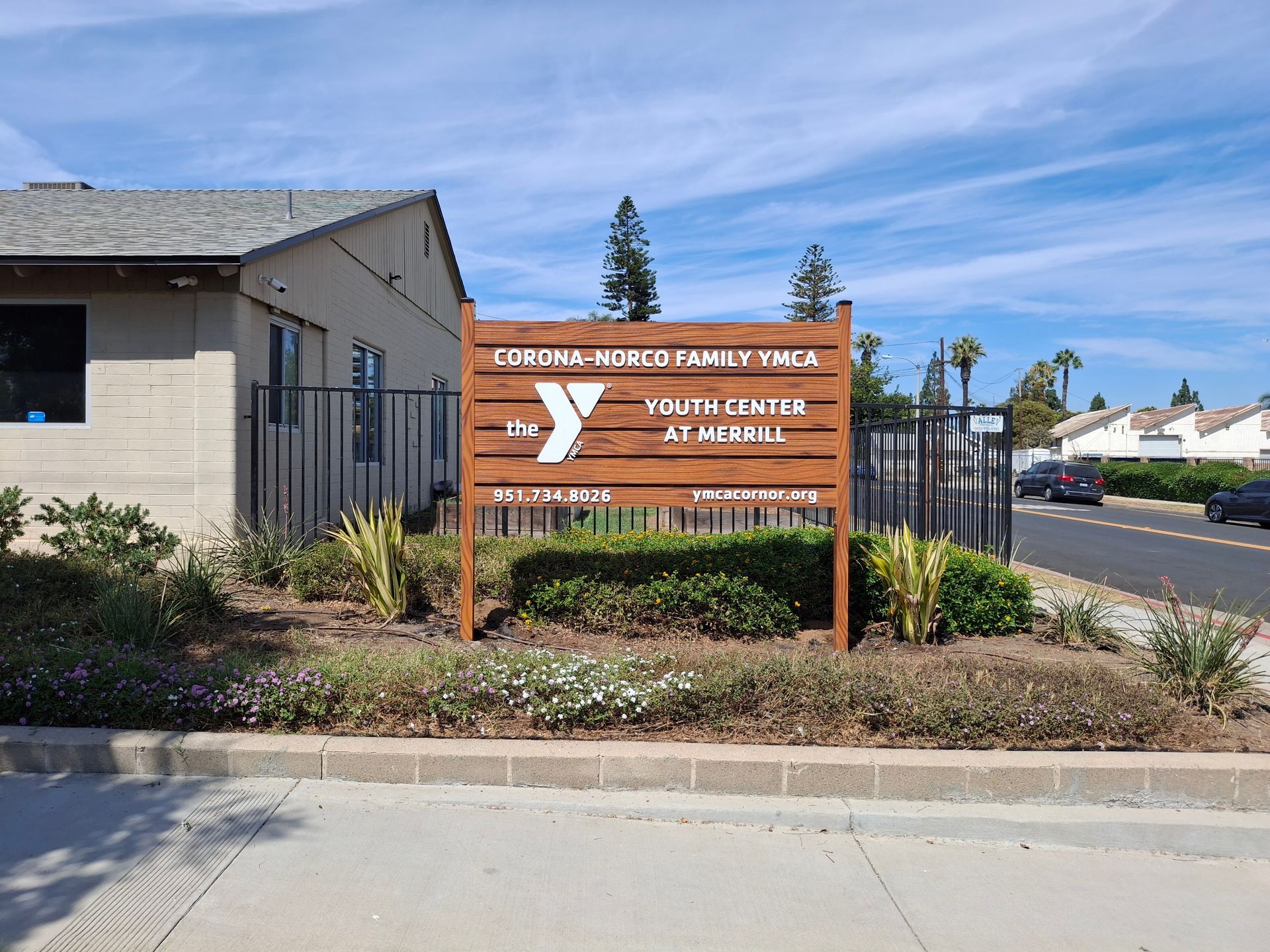 Wooden sign for YouthBuild at a street corner beside a beige building and landscaping.