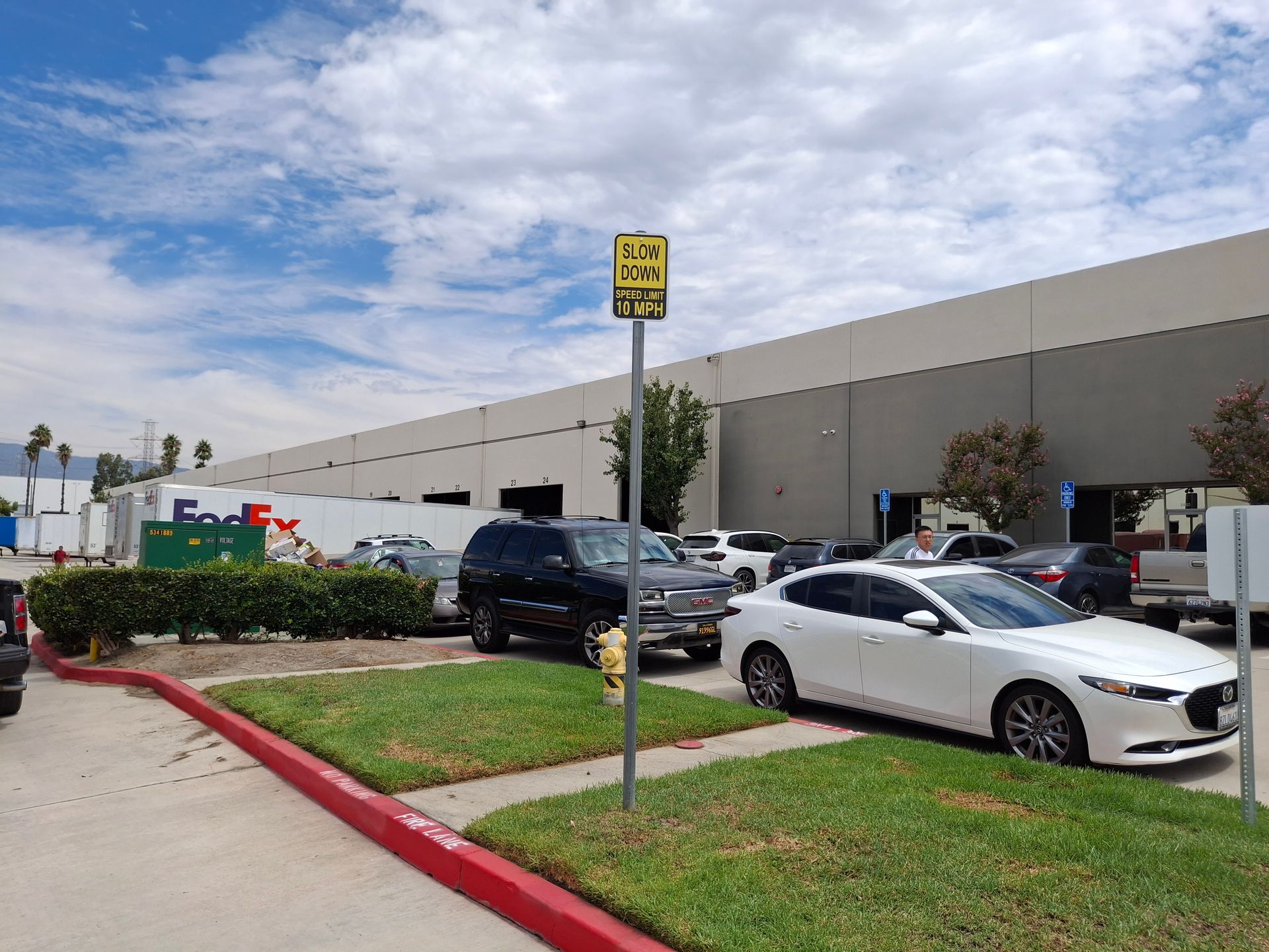 Parking lot with white sedan and black SUV beside a large warehouse under a cloudy sky.