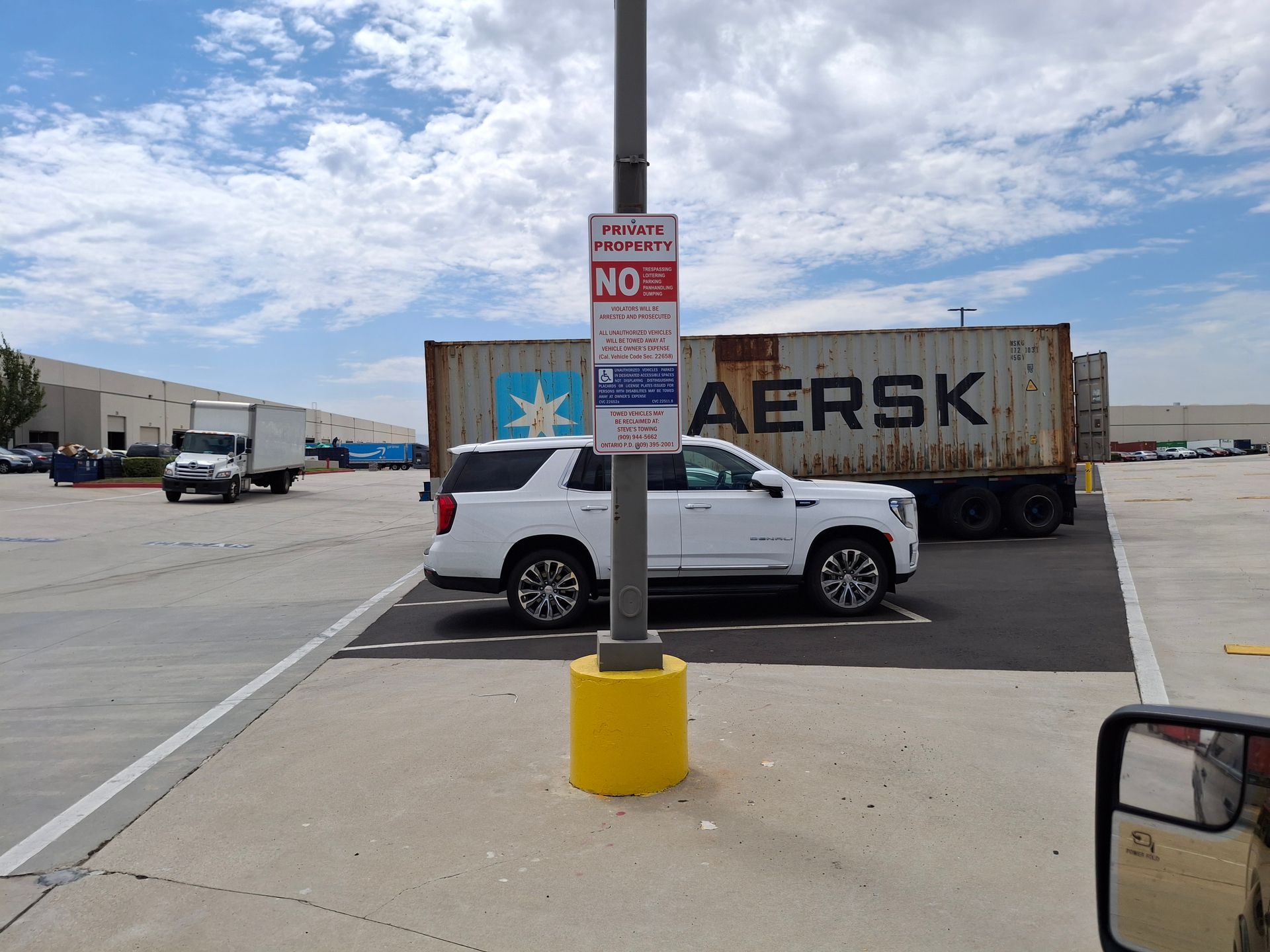 Parking lot with white SUV beside a Maersk trailer under a cloudy sky, viewed past a no-parking sign.
