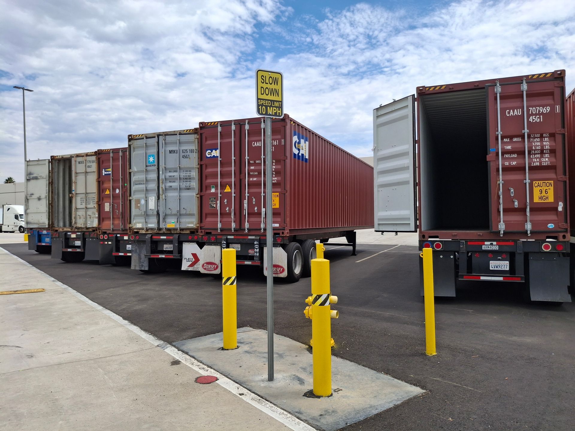 Trucks lined up by a warehouse loading dock with shipping containers and yellow safety posts.