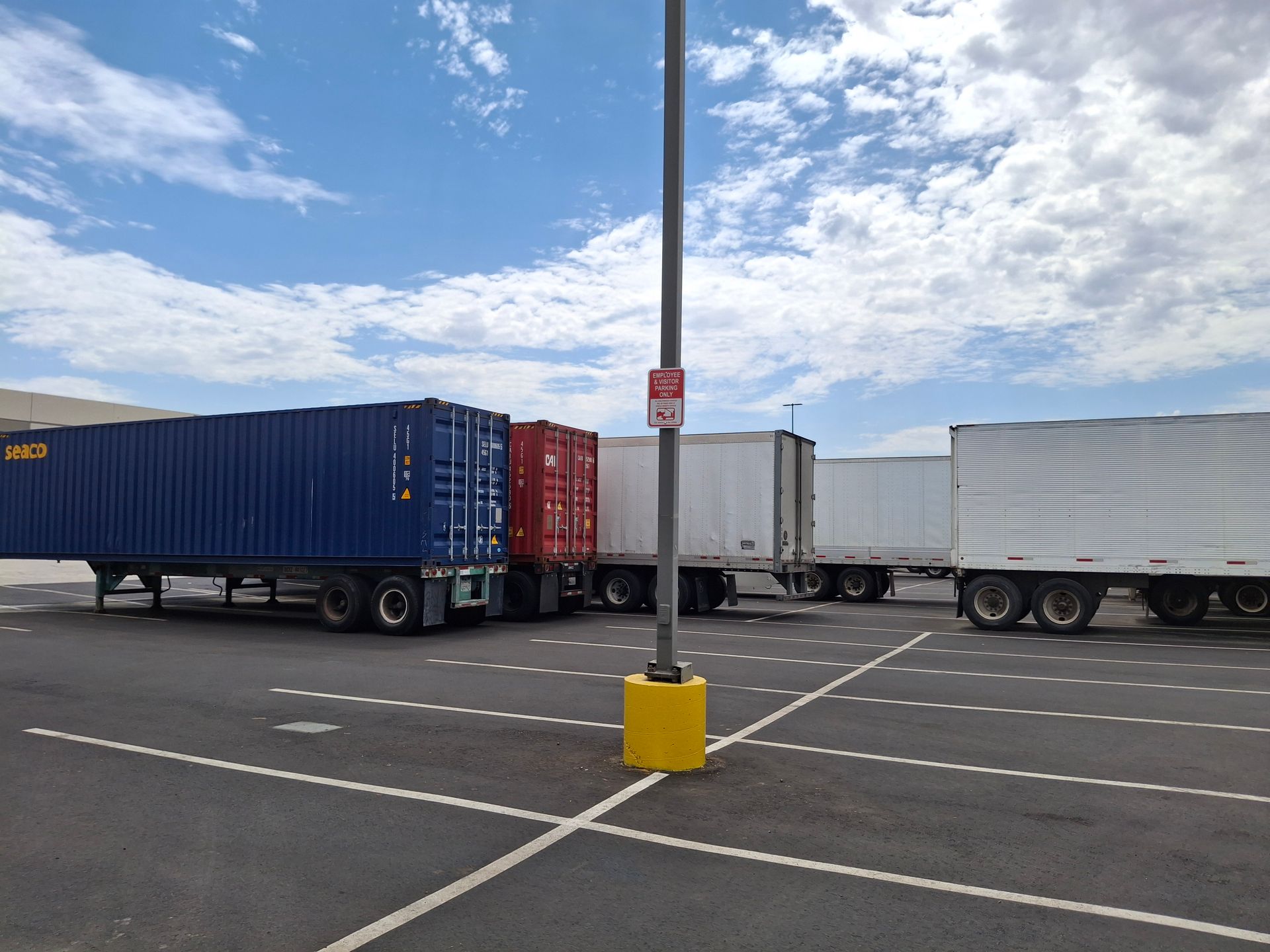 Semi-trailers parked in an empty lot beneath a cloudy blue sky, with a light pole in the center.