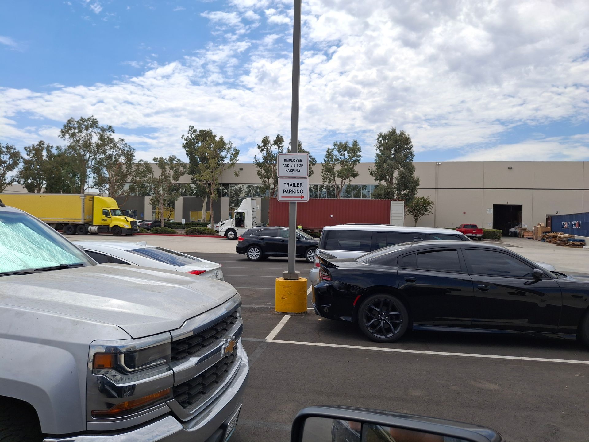 Parking lot with a black car, silver truck, and yellow truck near a warehouse under a cloudy sky