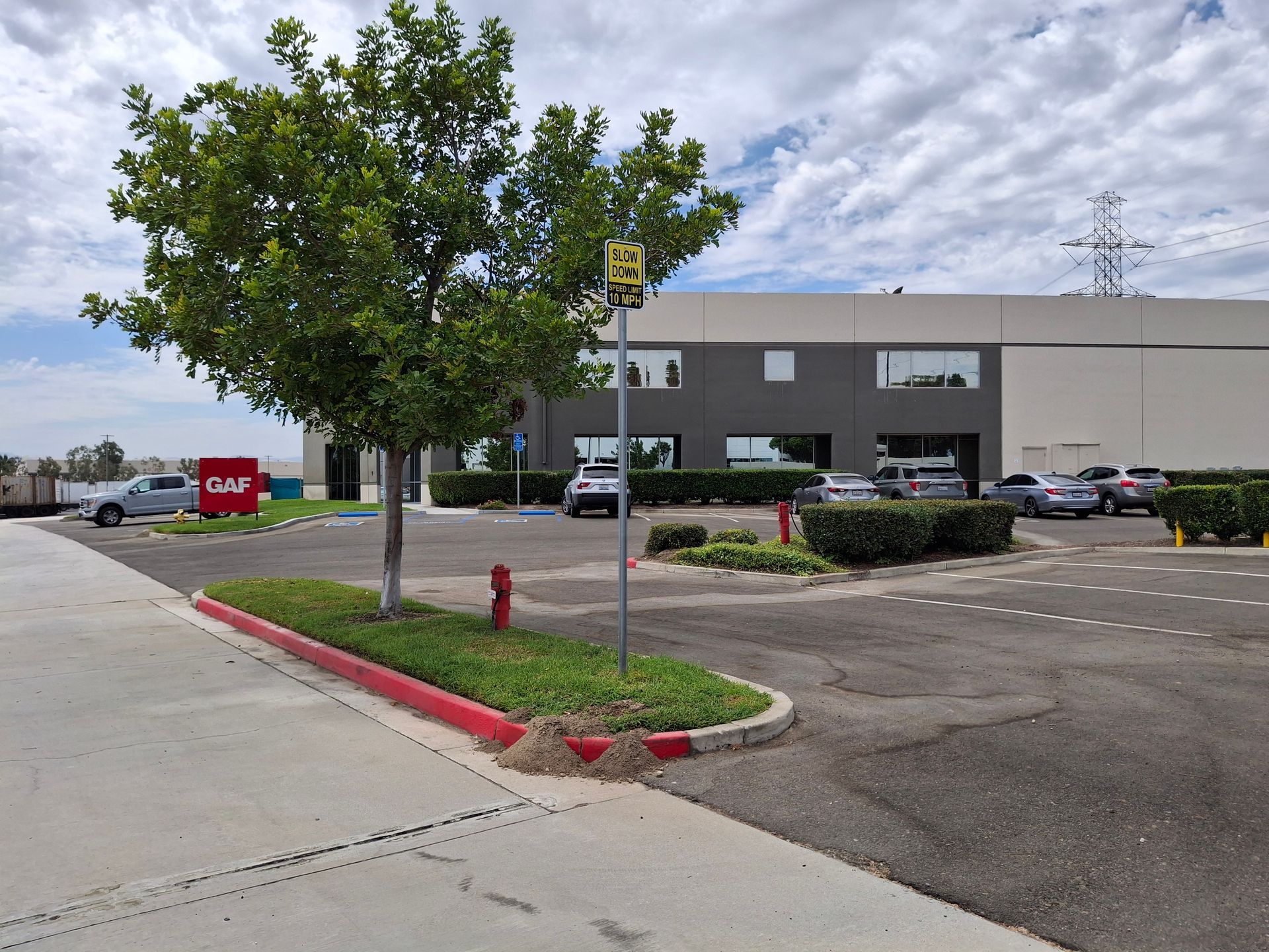 Parking lot beside gray commercial building with trees, parked cars, and a red curb in front