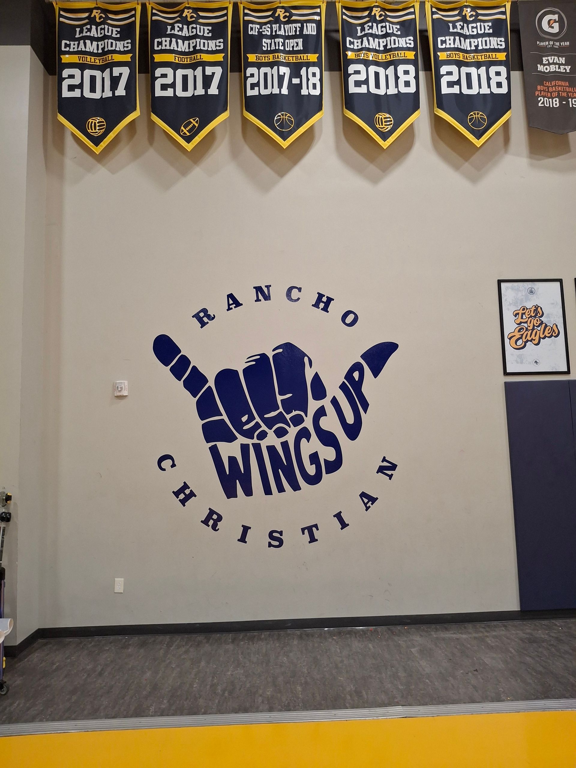 Rancho Christian wall logo in blue with championship banners above it in a gymnasium