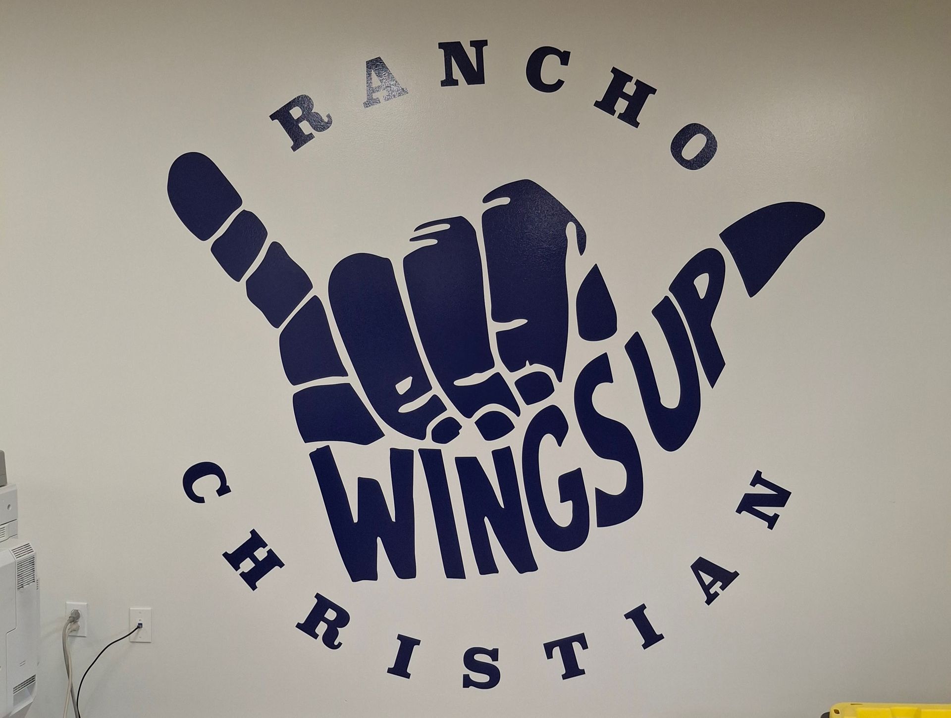 WingsUp Rancho Christian wall logo with a blue hand sign and circular text on a beige wall