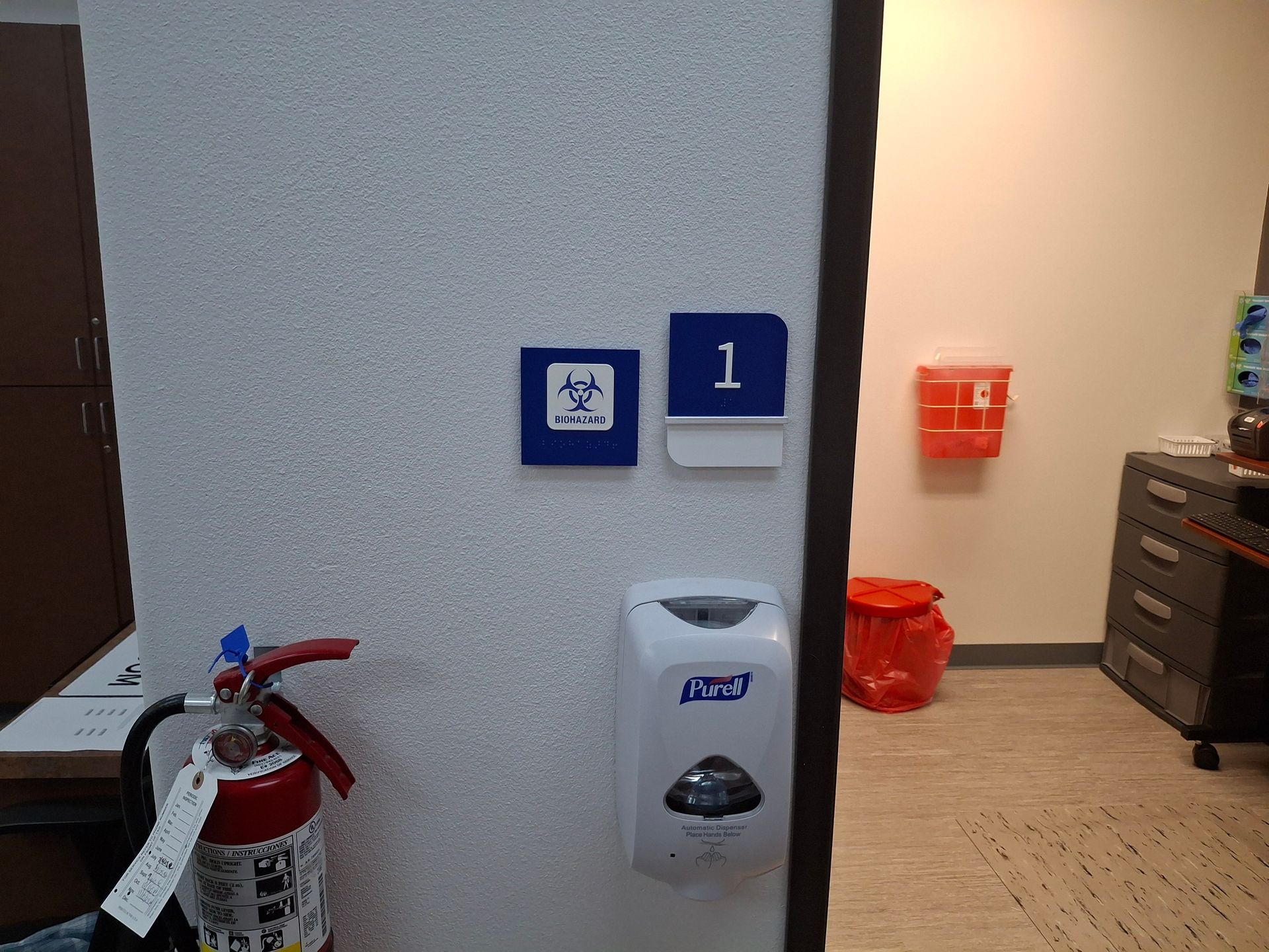 Office hallway with a fire extinguisher, sanitizer dispenser, and room number sign 1 beside a doorway