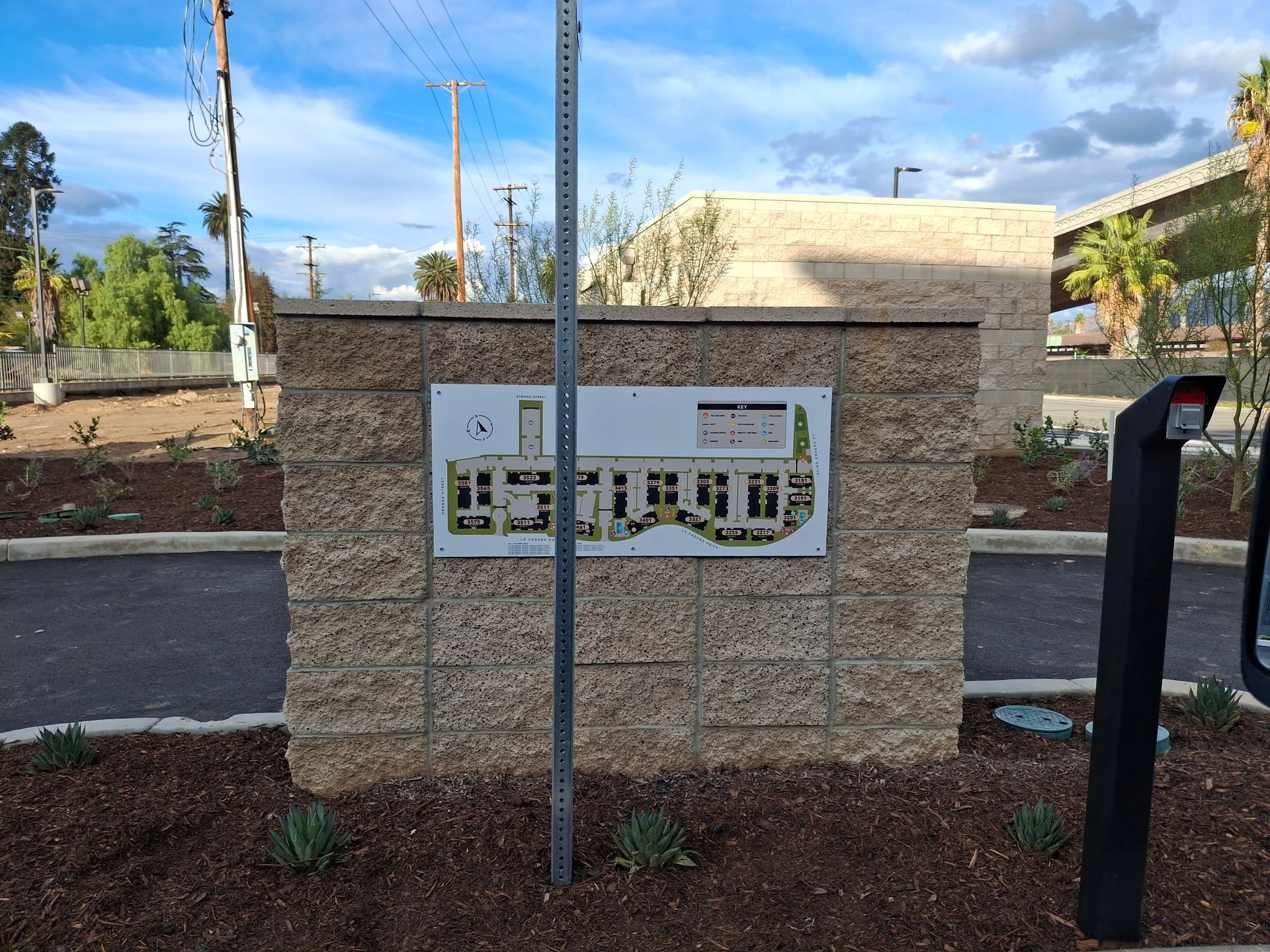 Apartment directory sign mounted on a tan stone wall beside a walkway and mailbox post