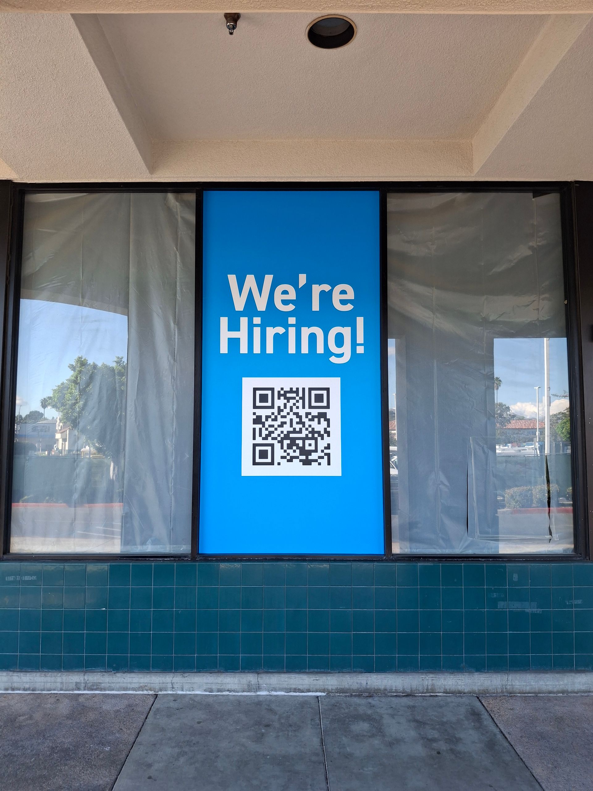 Storefront window with blue “We’re Hiring!” sign and QR code displayed in the center.