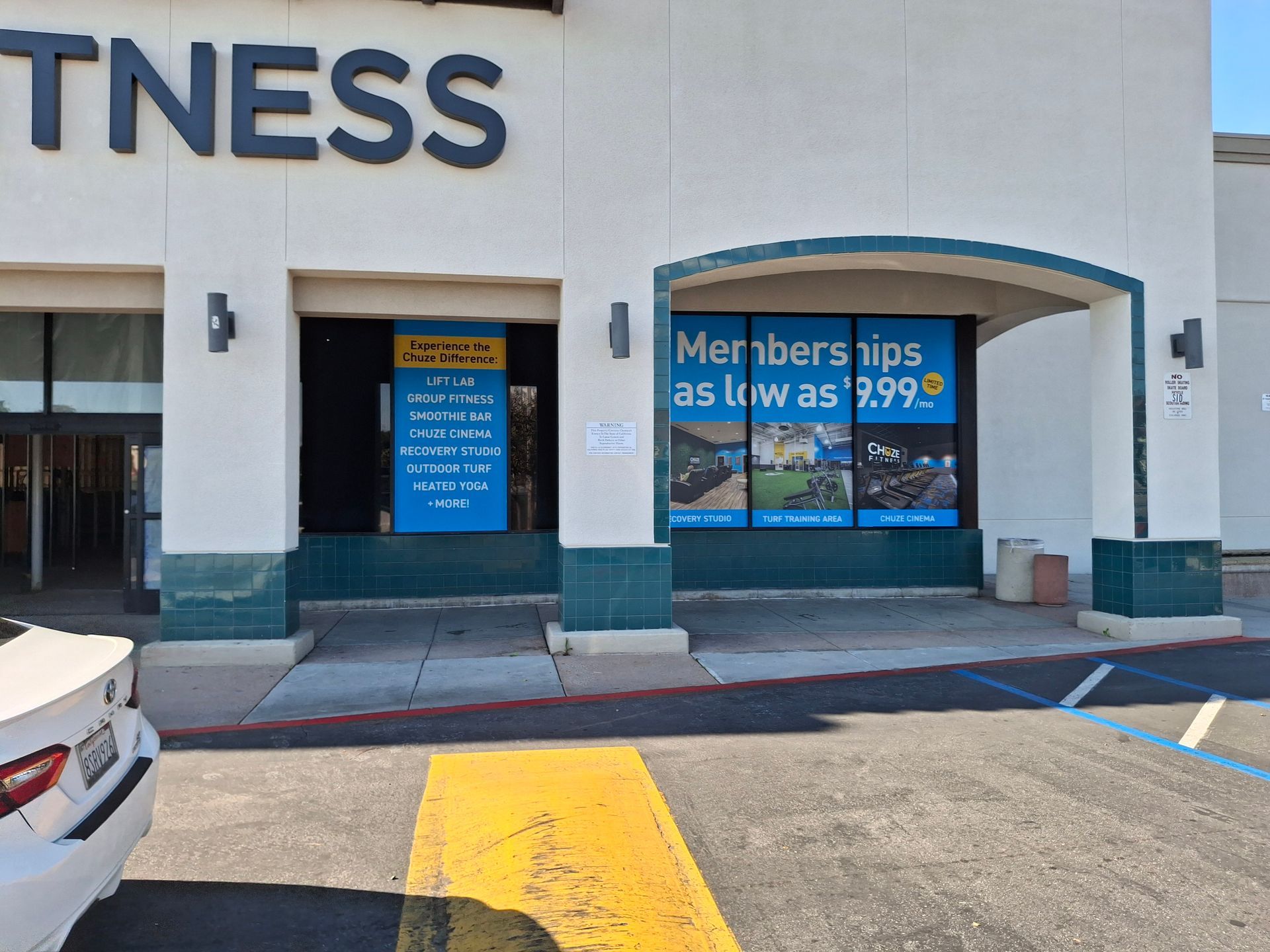 Gym storefront with blue doors, sign reading “Memberships as low as $19.99,” and parking lot in front