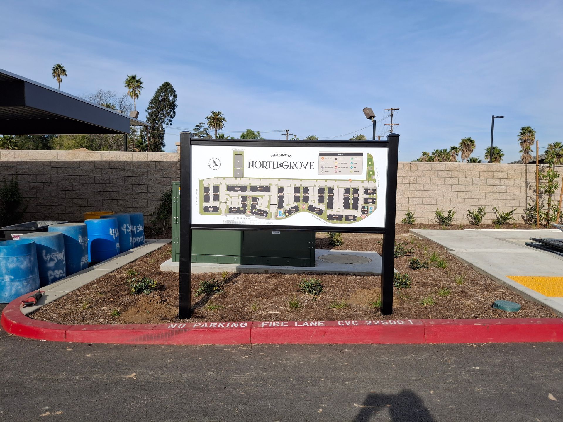 Outdoor park signboard on a landscaped median with red curb, stone wall, and blue bins nearby
